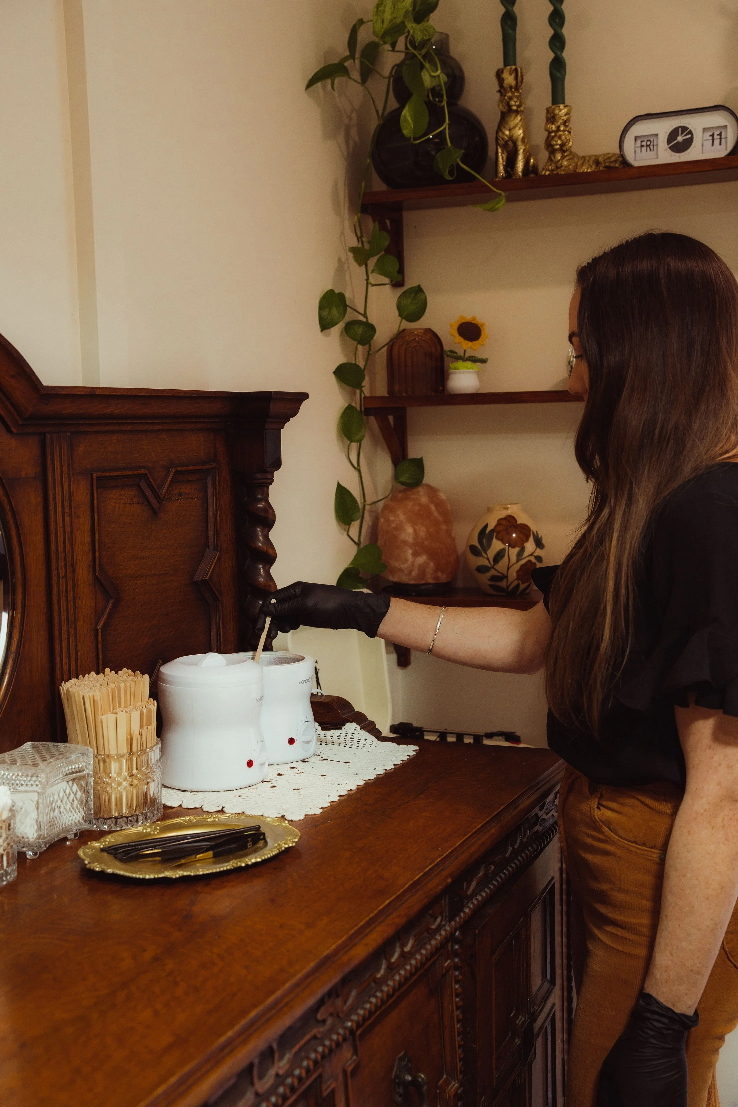 A woman wearing black gloves is filling a hot water dispenser on a wooden sideboard in a home setting.