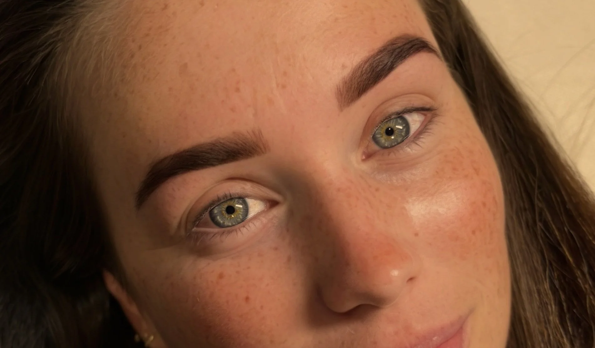 Close-up of a woman's face with tattooed eyebrows, clear skin, and light-colored eyes, lying down and looking upward.