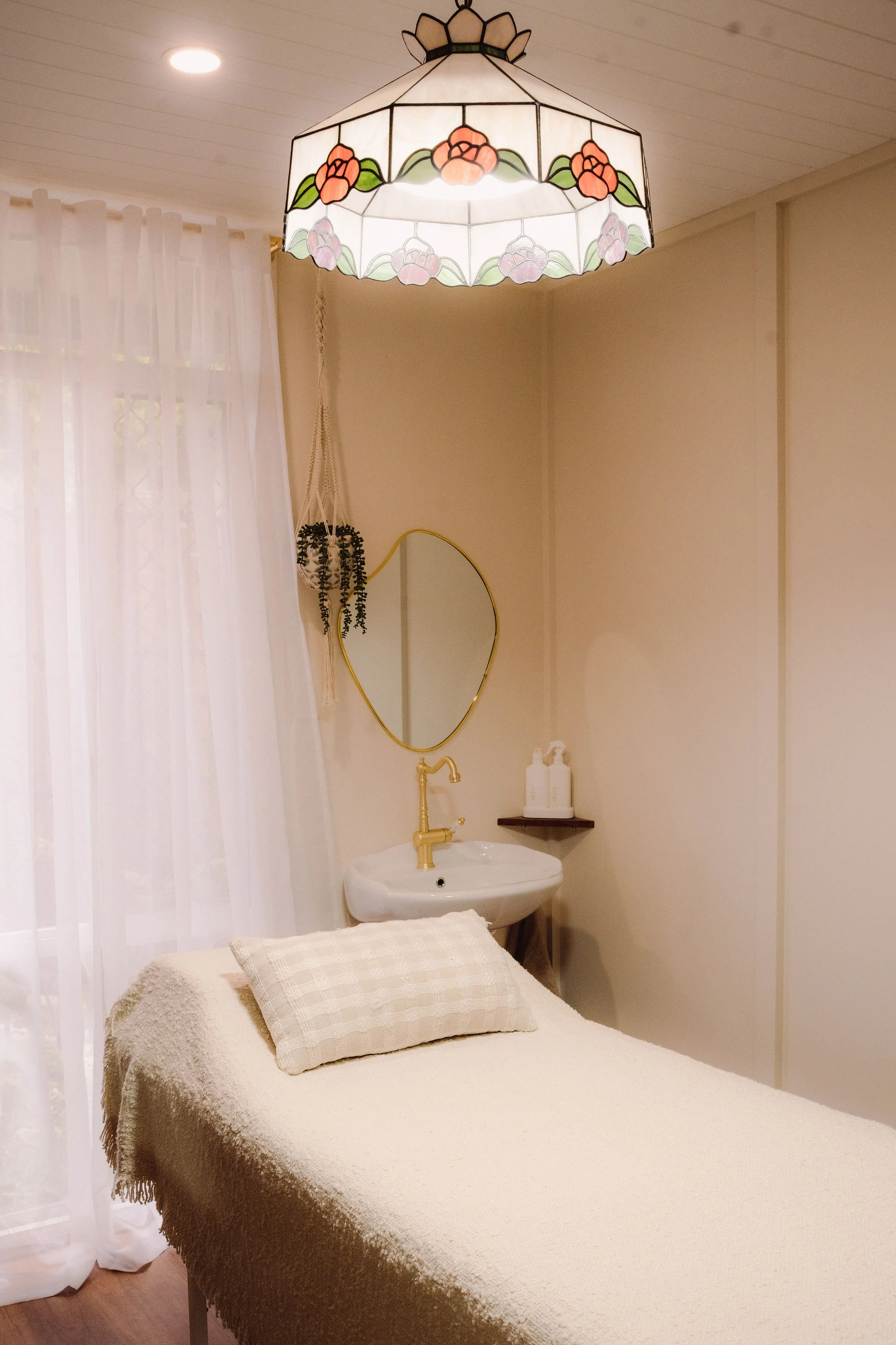 A cozy room with a massage table, a pendant lamp with floral stained glass design, sheer curtains, a small sink with a mirror, and bottles on a shelf.