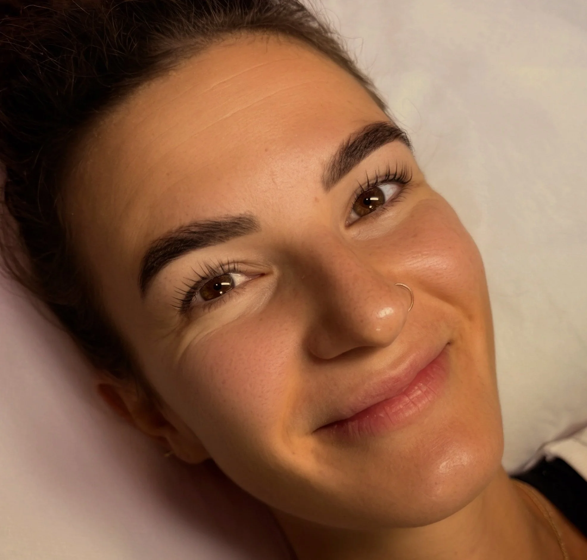 Close-up of a smiling woman with brown eyes, dark eyebrows, freckles, a small nose ring, and long eyelashes.