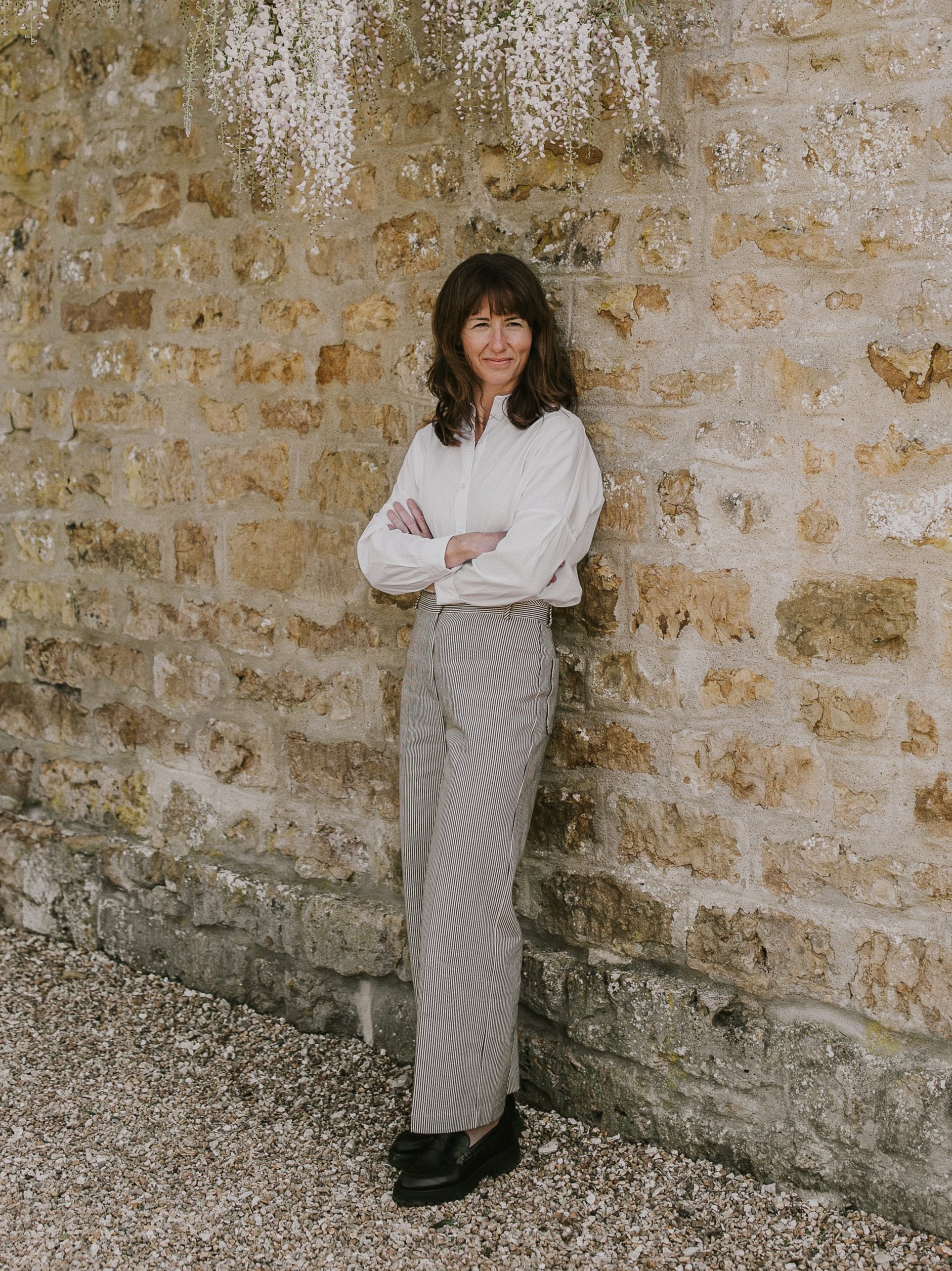 Studio RDC Limited — Studio RDC. Ruth Chadney. Architect. A woman with dark brown hair in a white blouse and striped trousers leaning against a rustic stone wall with hang