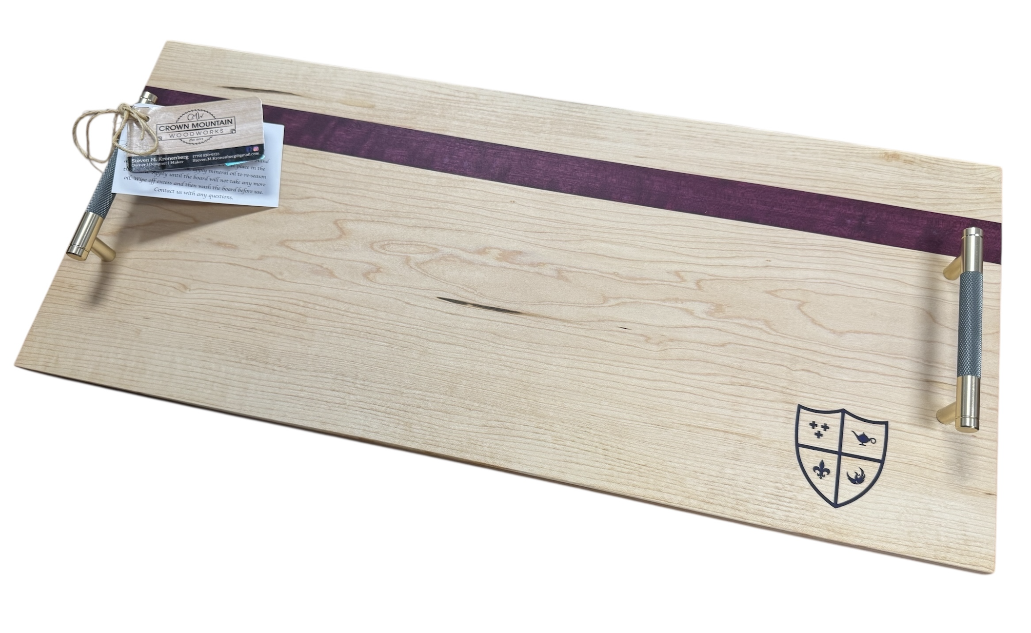 A rectangular wooden tray with metal handles on each side, featuring a purple stripe across the top. There is a small tag attached to the left handle, and a shield-shaped logo with four quadrants on the lower right corner of the tray.