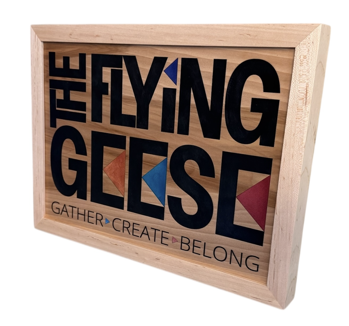 Decorative wooden sign with the words "The Flying Geese" and the phrase "Gather, Create, Belong" with colorful geometric bird shapes.