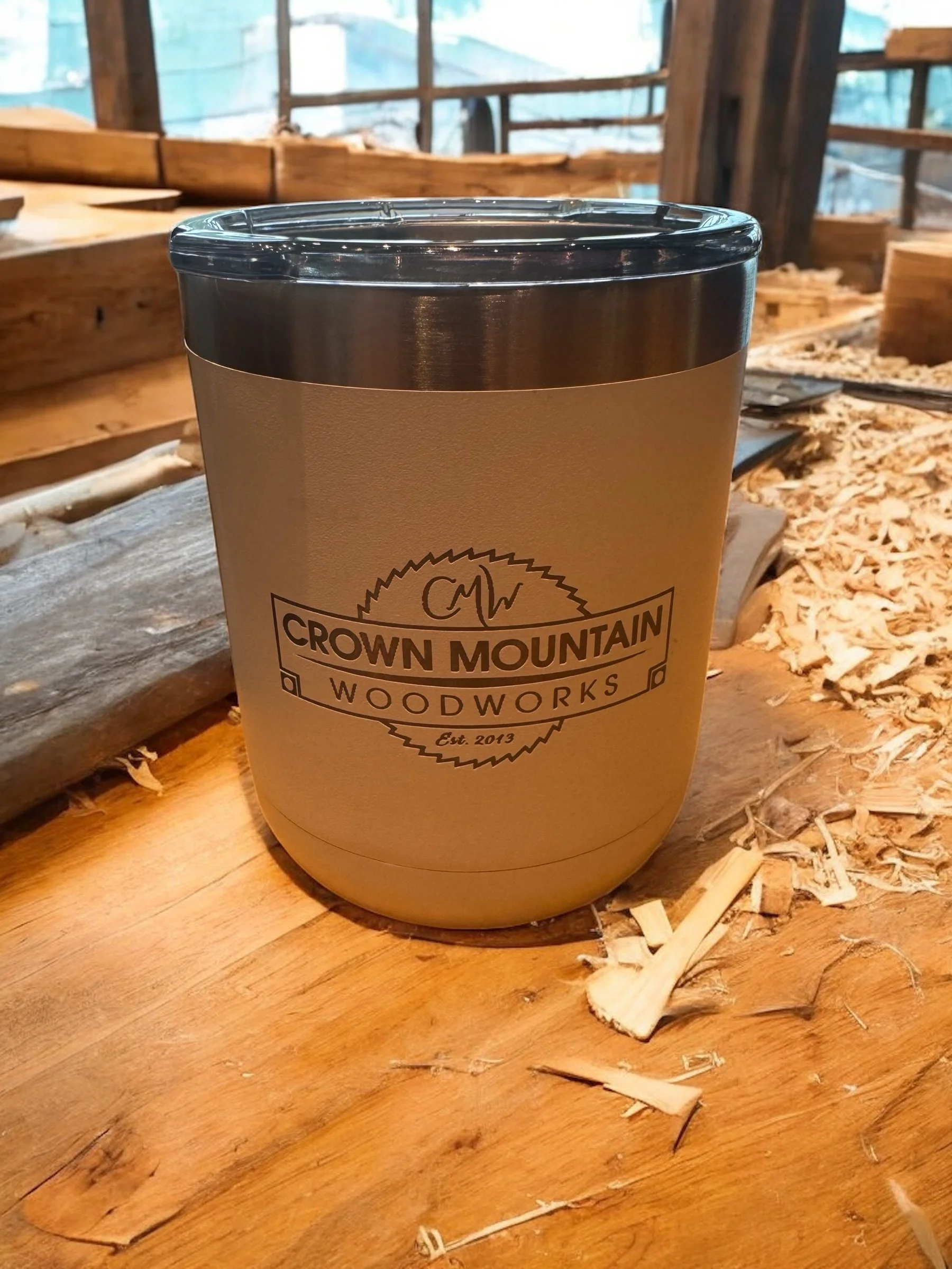 A metal insulated tumbler with a beige exterior and lid on a wooden workbench, with wood shavings and tools in the background, logo reads 'Crown Mountain Woodworks' and 'Est. 2013'.