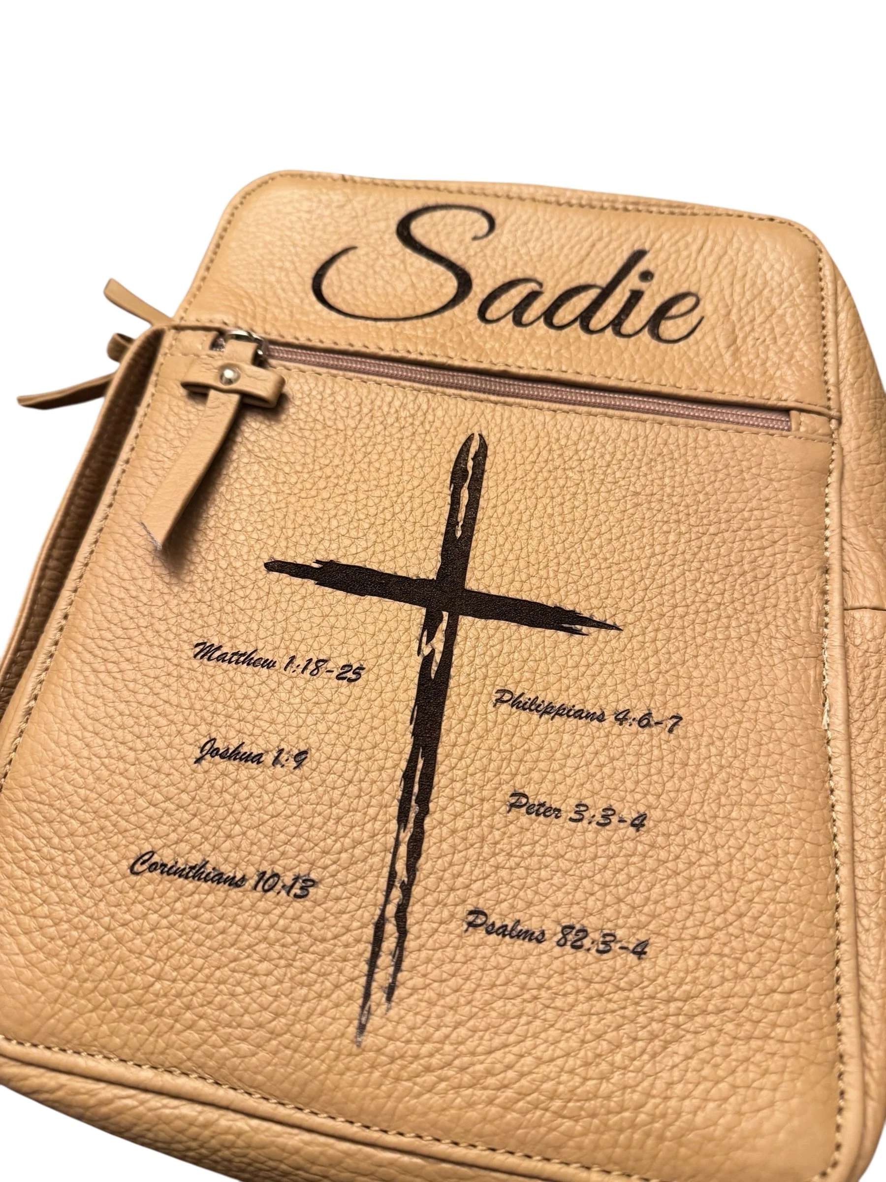 Tan leather backpack with office-style zipper, decorated with a black cross and Bible verse references.