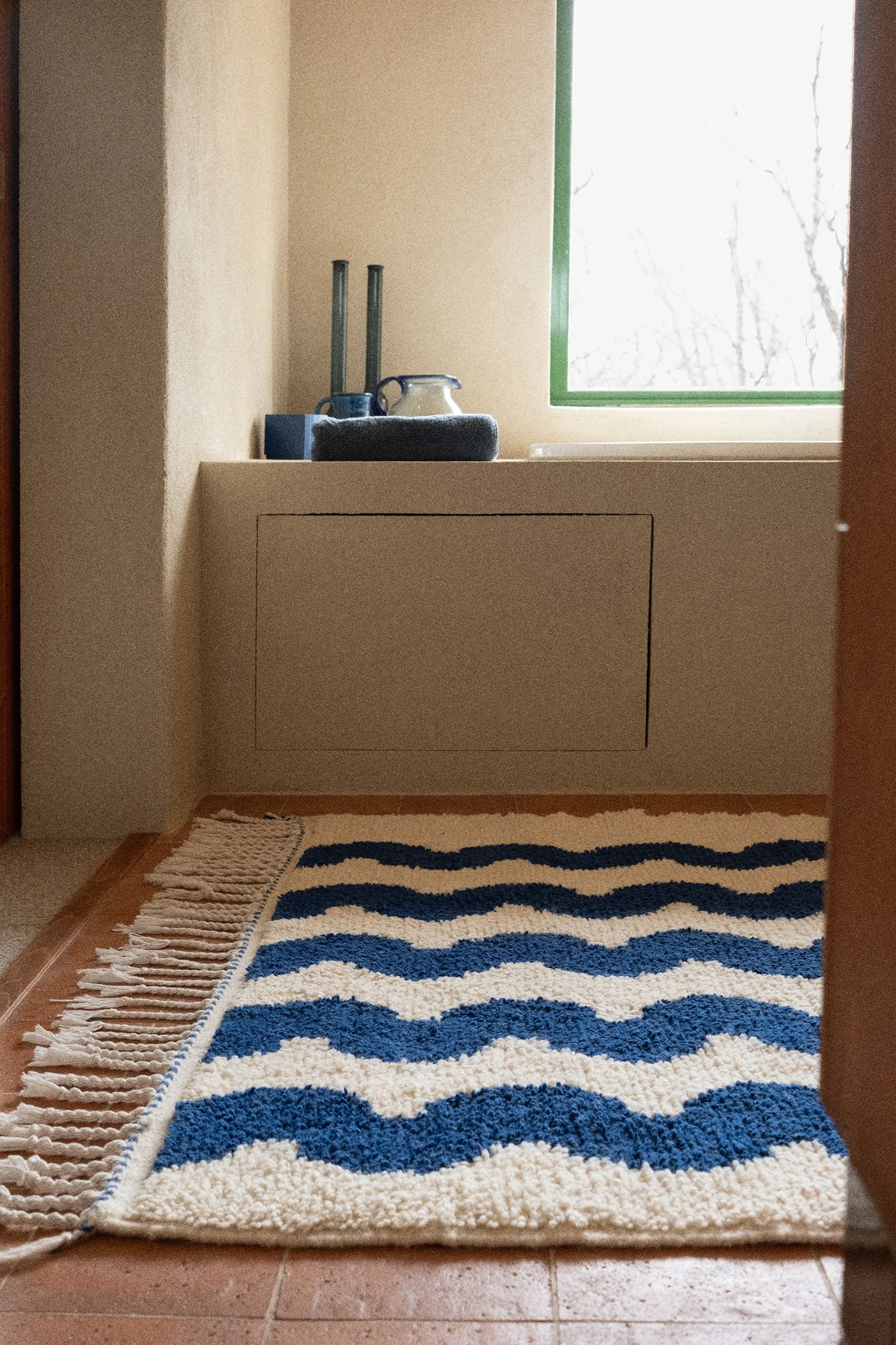 Bathroom corner with a terracotta floor, a blue and white wavy-patterned Karpet rug, and a ledge adorned with candles and a ceramic pitcher.