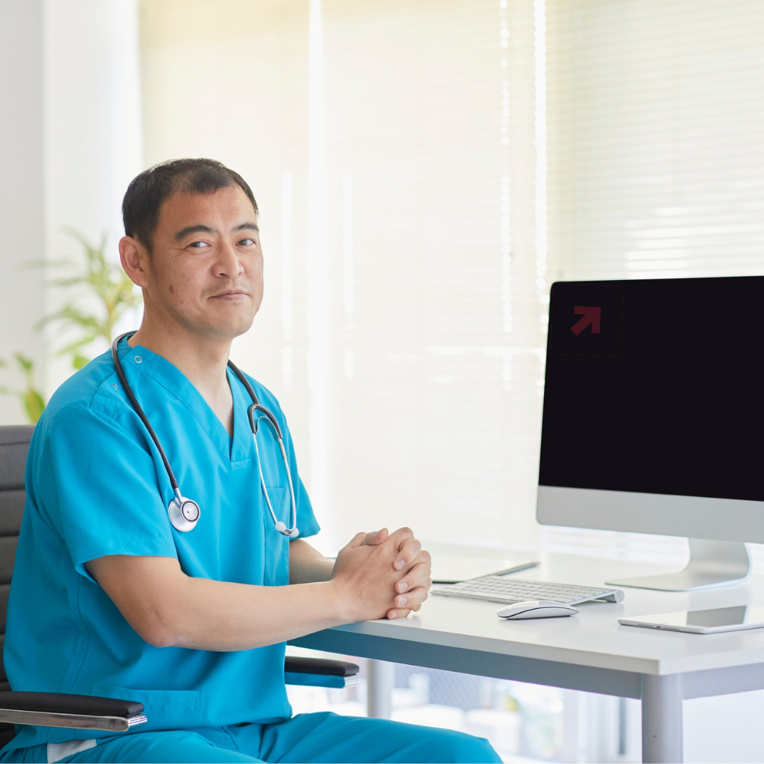 A male healthcare worker in blue scrubs with a stethoscope around his neck sits at a white desk next to a computer in a bright room.