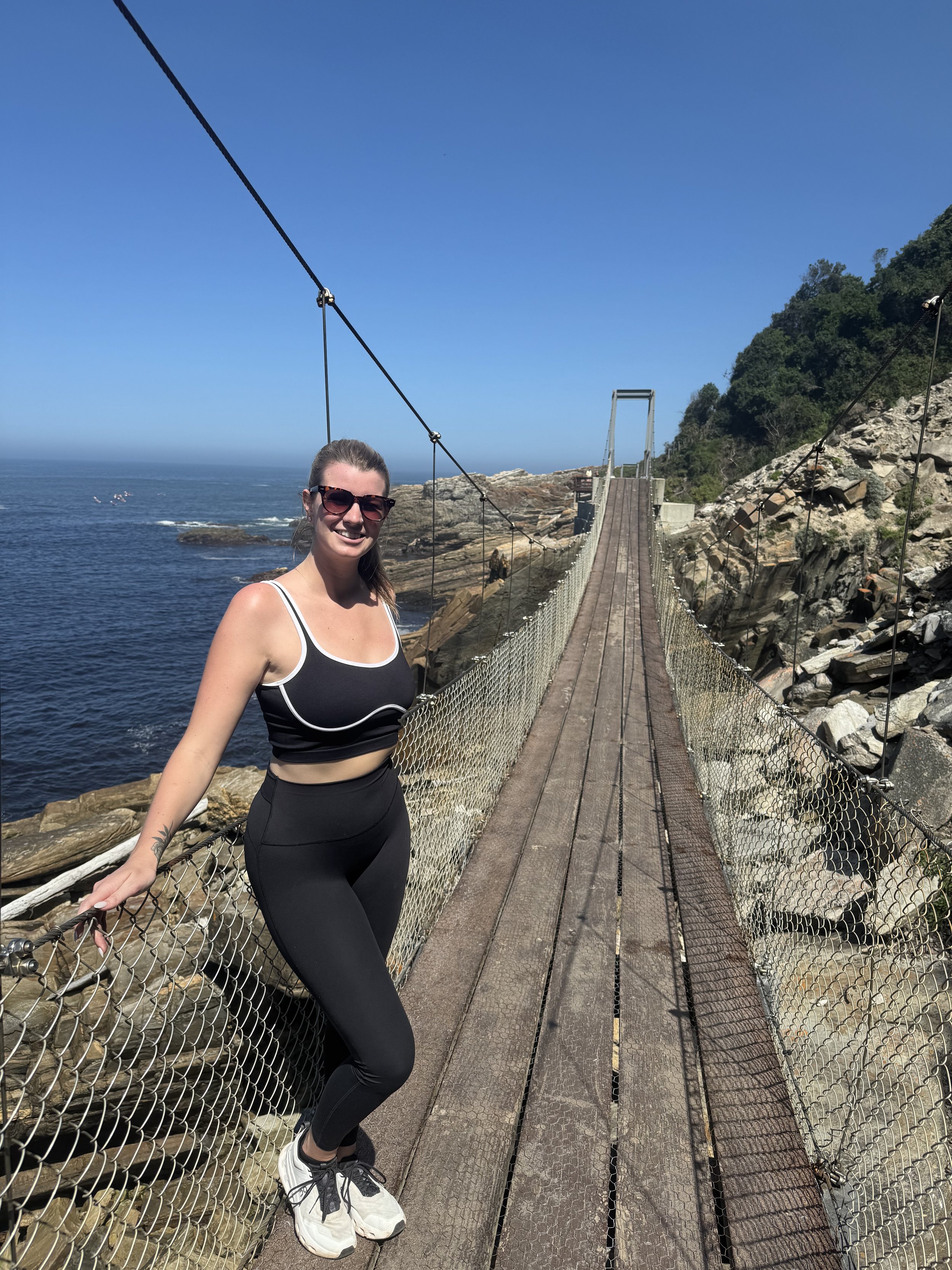 Woman standing on a suspension bridge over water, wearing athletic attire and sunglasses, with rocky shoreline and blue sky in the background.