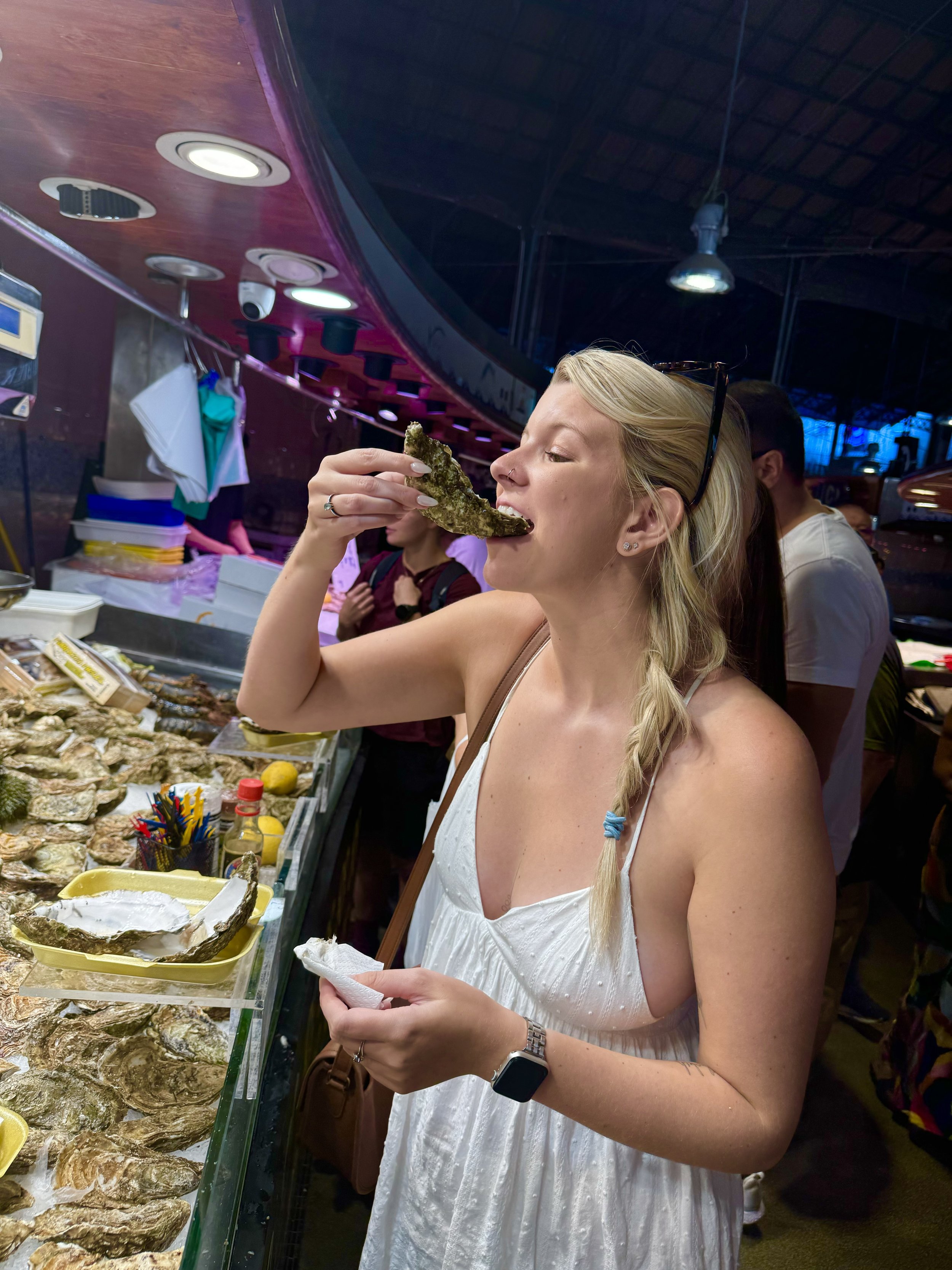 Woman in a white dress eating an oyster at a seafood market.