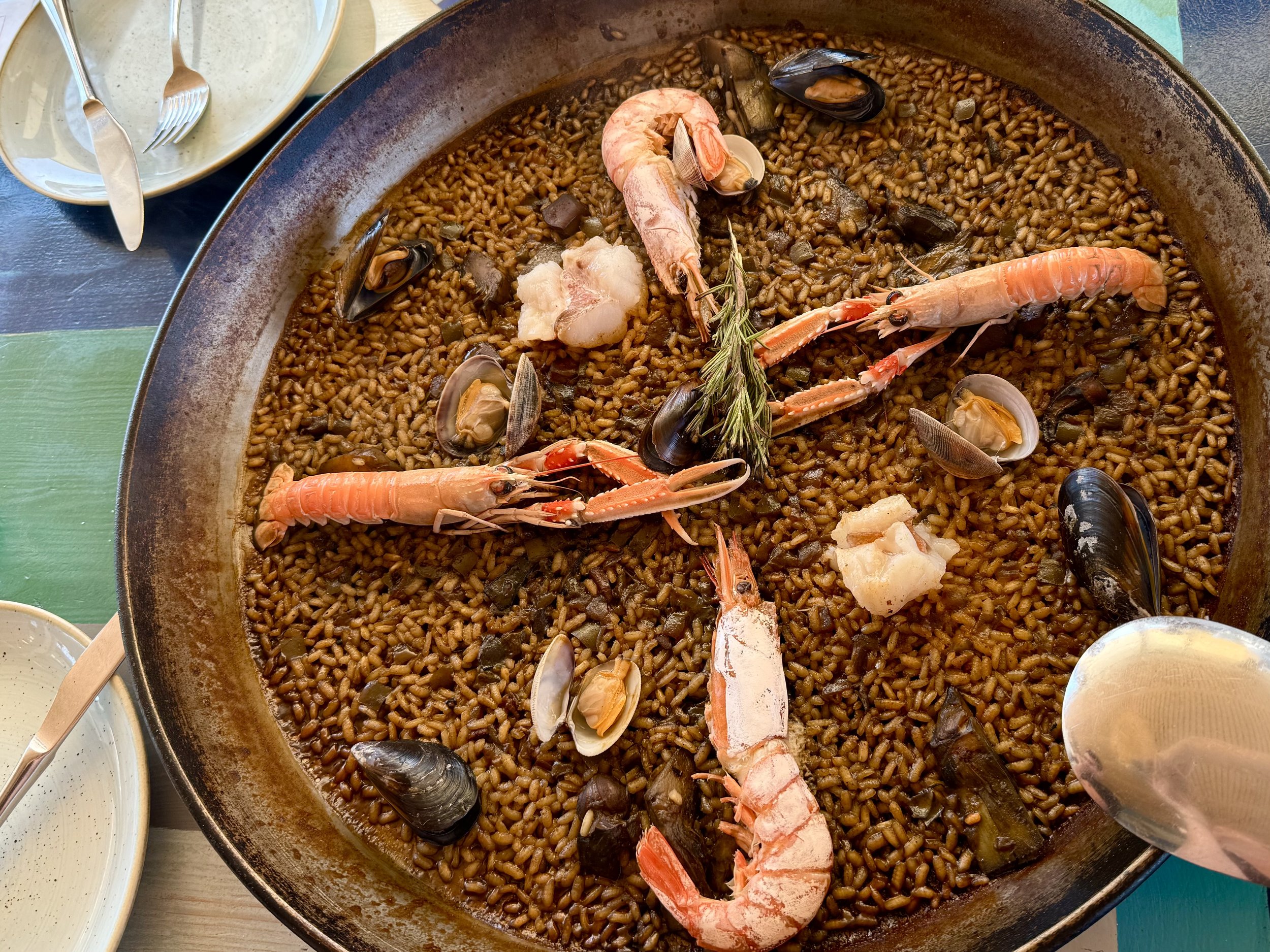 Seafood paella with shrimp, mussels, clams, and herbs in a large pan on a table with cutlery and plates nearby.