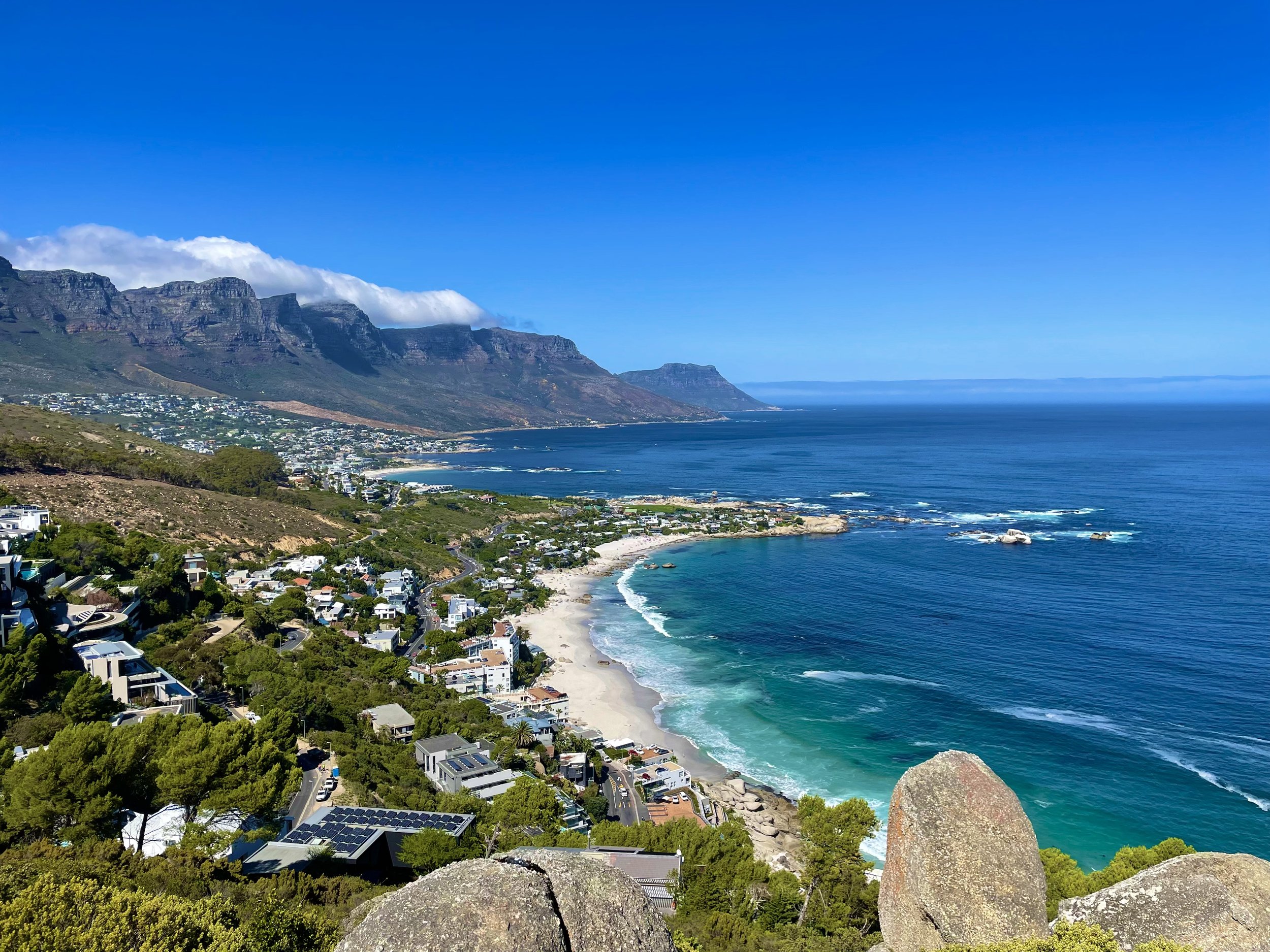 Coastal view of a seaside city with mountains, houses, and a sandy beach under a blue sky in Cape Town, South Africa.