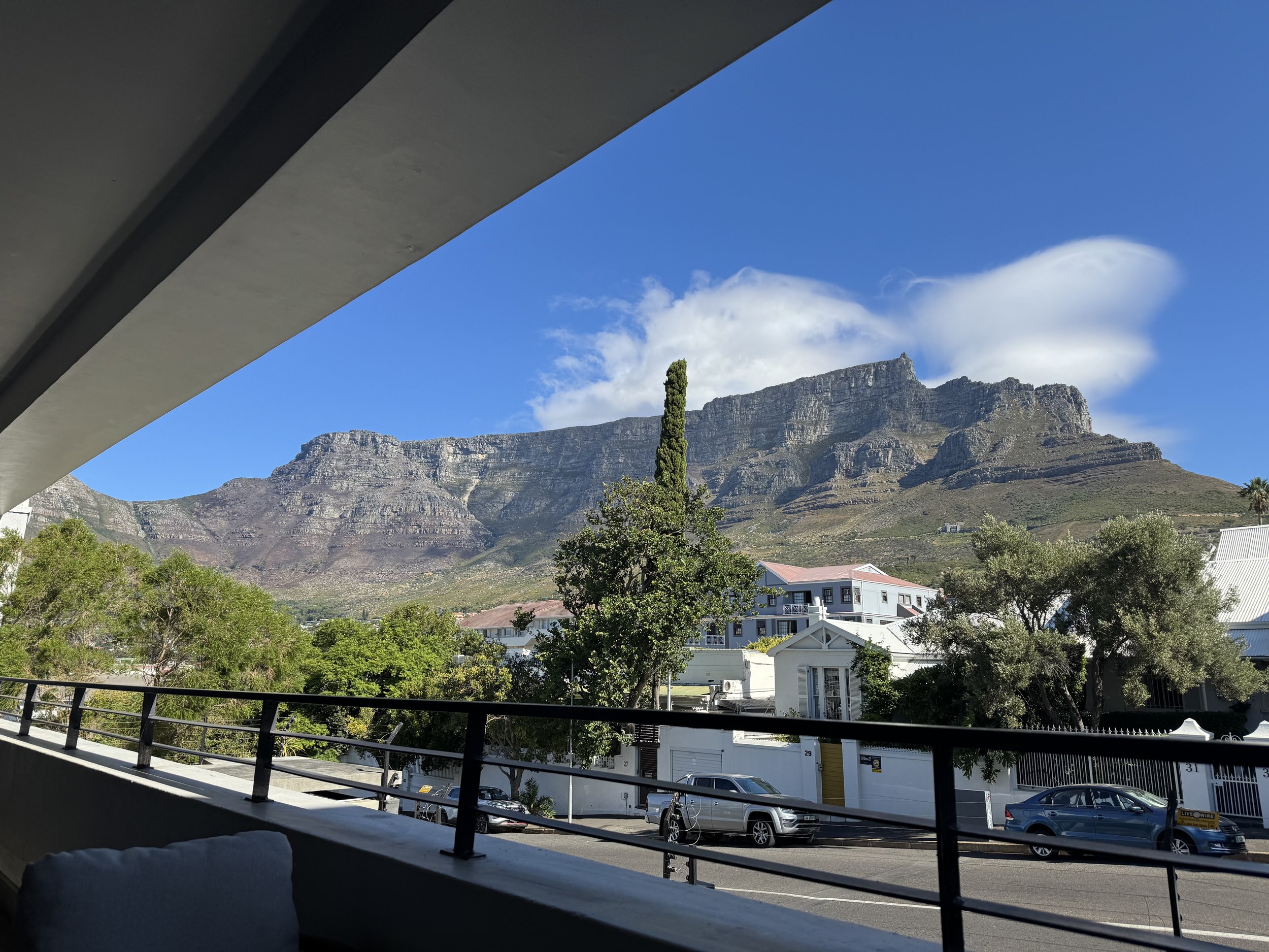 View of Table Mountain and surrounding landscape from a balcony with railings, with residential buildings and parked cars below, under a clear blue sky with some clouds.