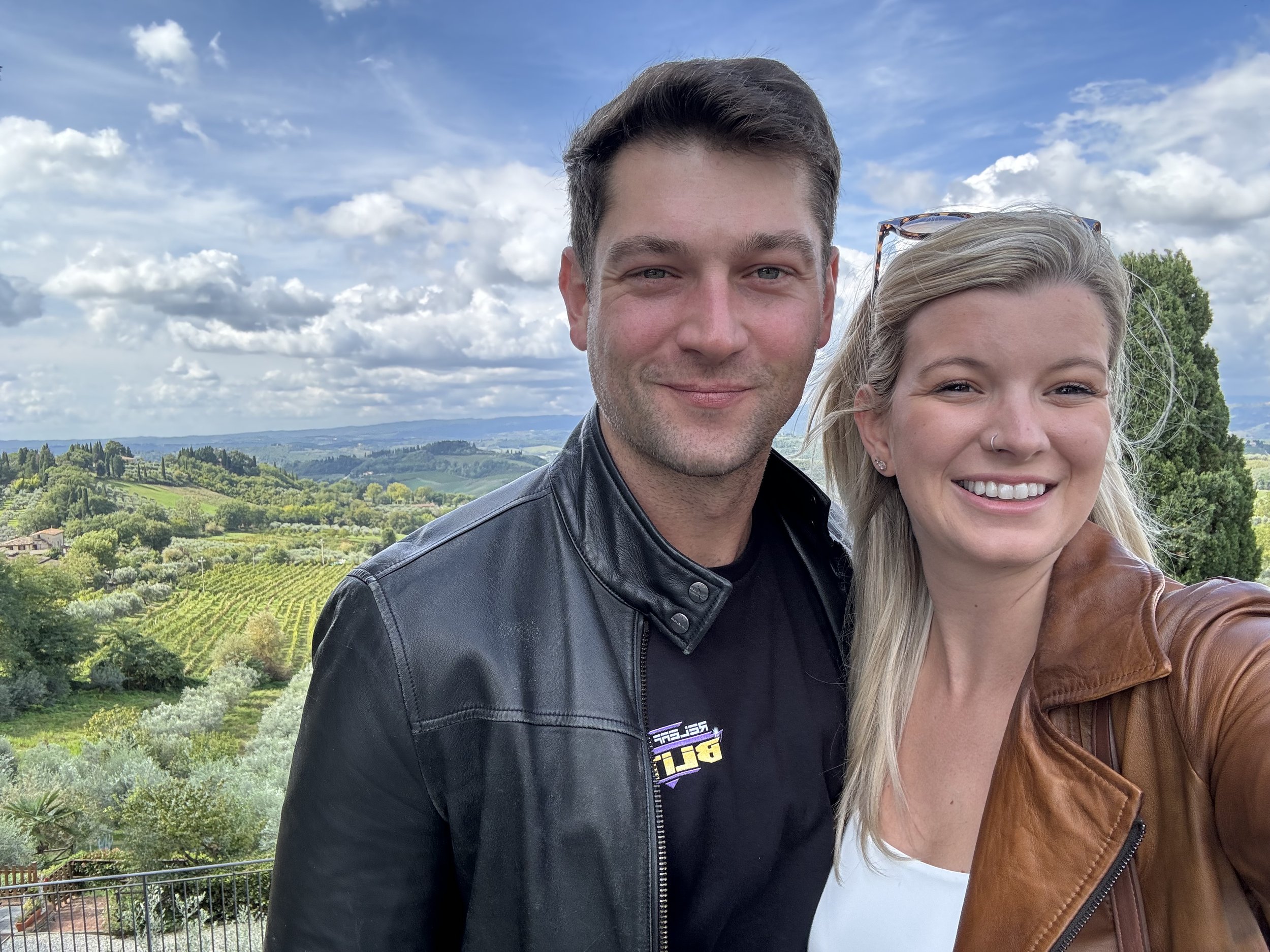 Two people taking a selfie with a scenic landscape of green hills and cloudy sky in the background.