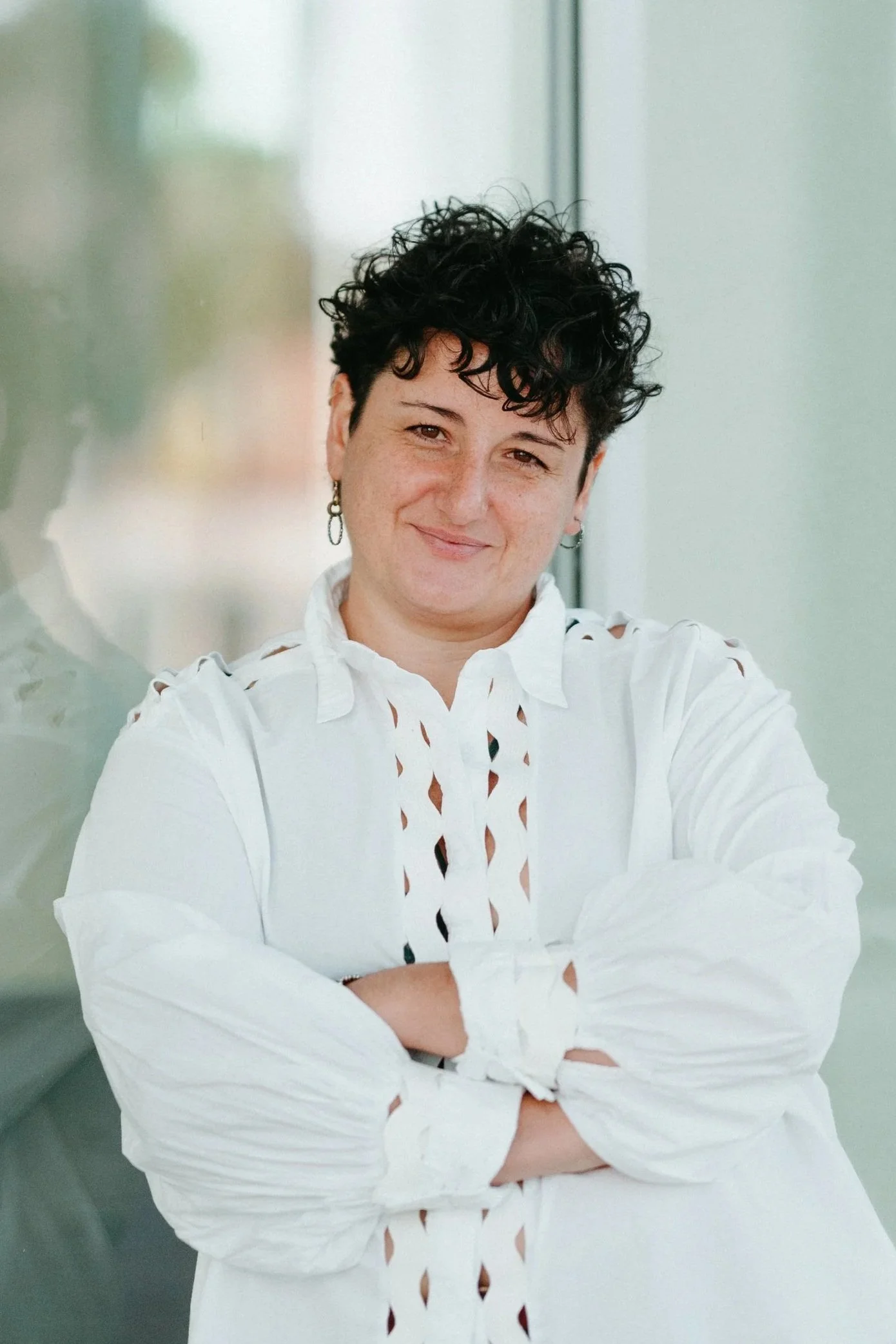A woman with short curly dark hair, wearing a white shirt with cutout patterns, crossing her arms and smiling.
