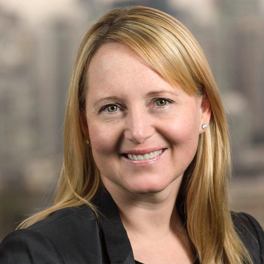 A woman with straight, shoulder-length blonde hair, wearing small earrings and a black blazer, smiling for a portrait against a blurred background.