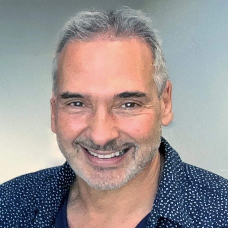 Close-up photo of a smiling middle-aged man with gray hair and beard, wearing a dark patterned shirt.
