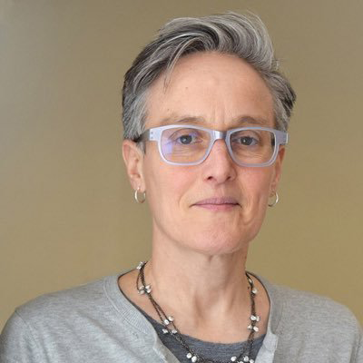 A middle-aged person with short gray hair and glasses, wearing a light gray shirt and a necklace, standing against a plain beige background.