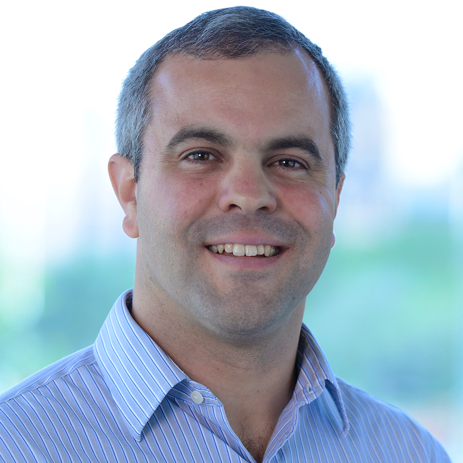 Close-up of a smiling man with short dark hair wearing a blue striped dress shirt, outdoors with blurred background of trees and sky.