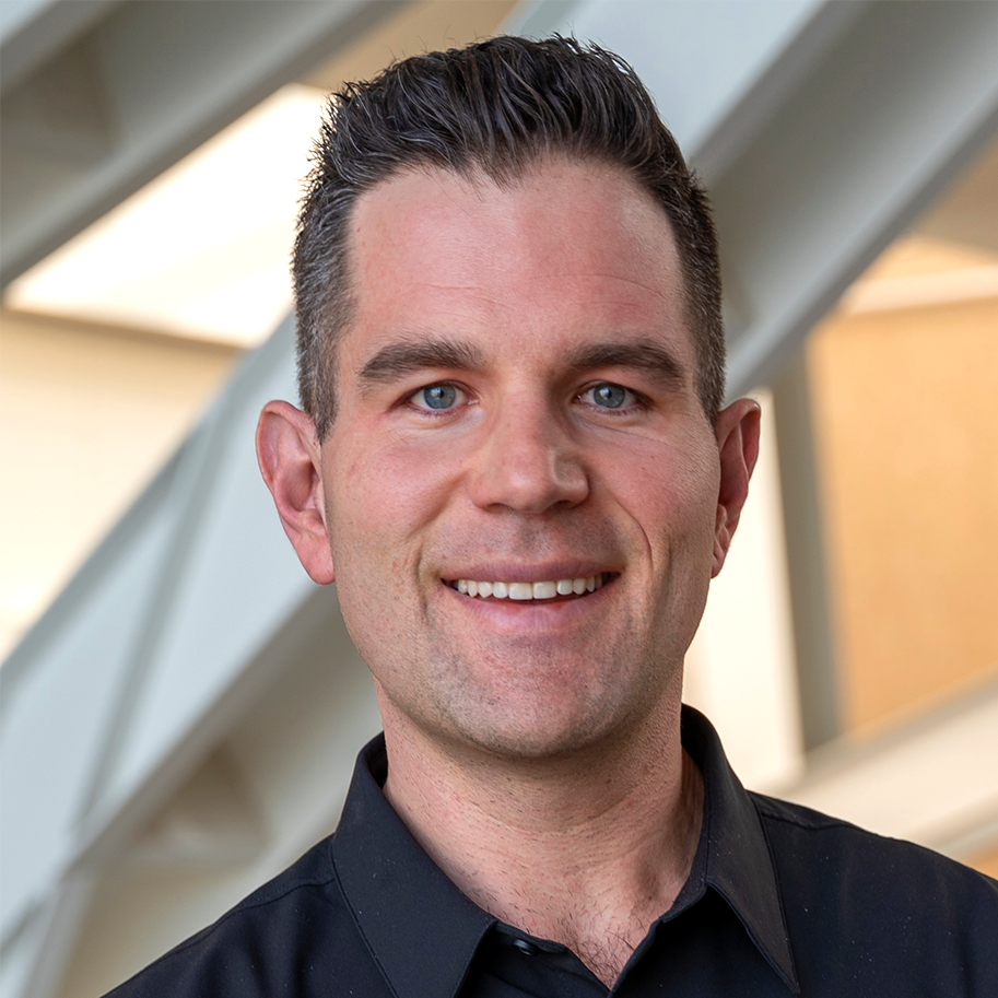Close-up of a smiling man with dark hair and blue eyes, wearing a black collared shirt, indoors with a modern architectural background.