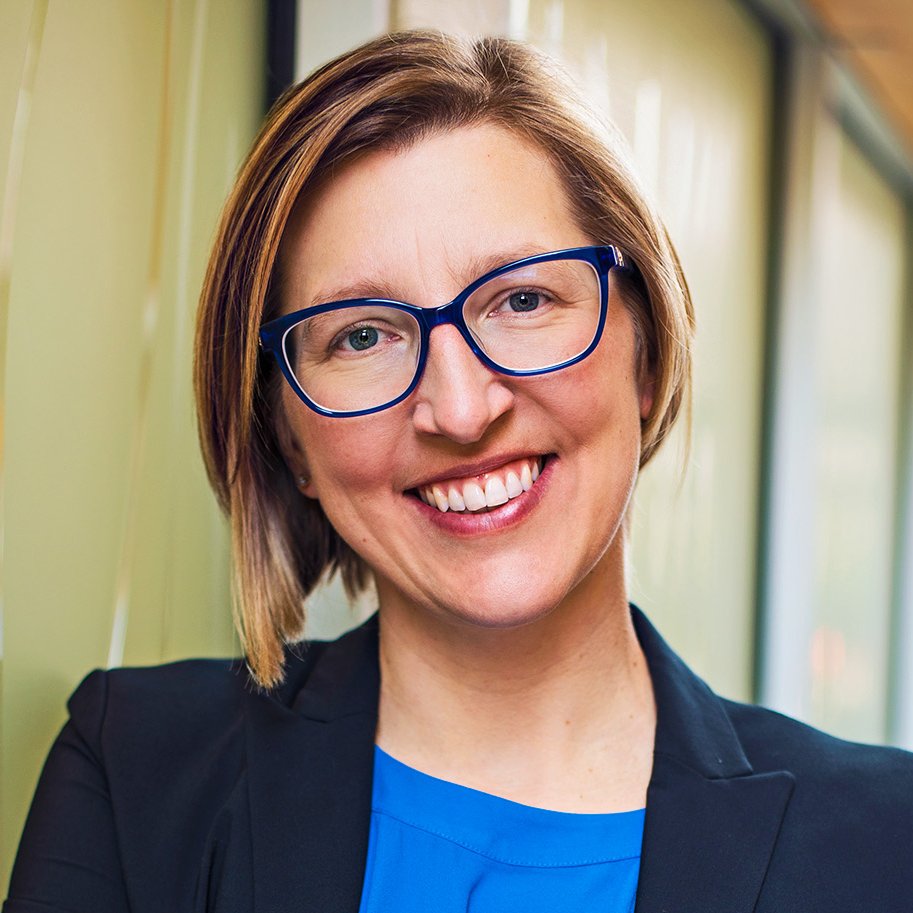 A woman with short, light brown hair, wearing blue glasses, a black blazer, and a blue top, smiling in an indoor setting with large windows.