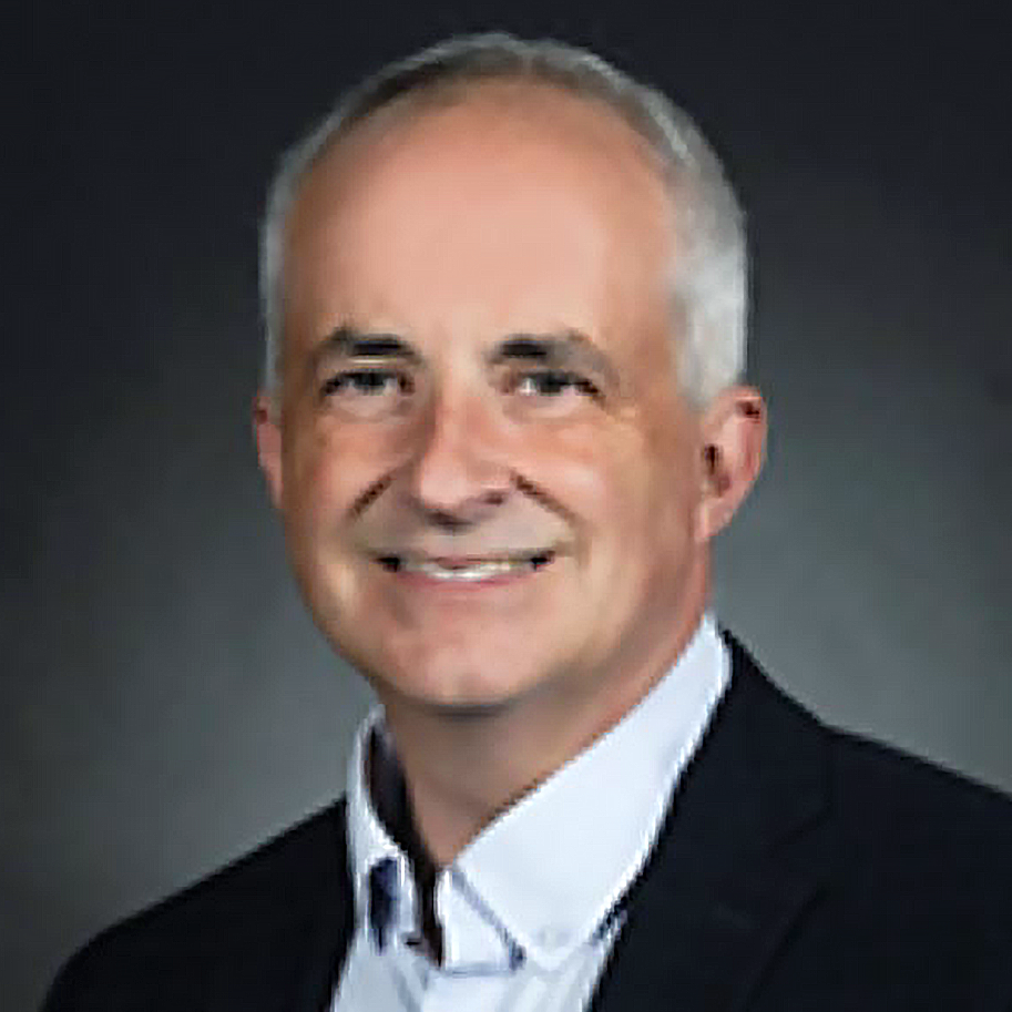A professional headshot of a middle-aged man with short gray hair, wearing a dark suit and white shirt, smiling against a dark background.