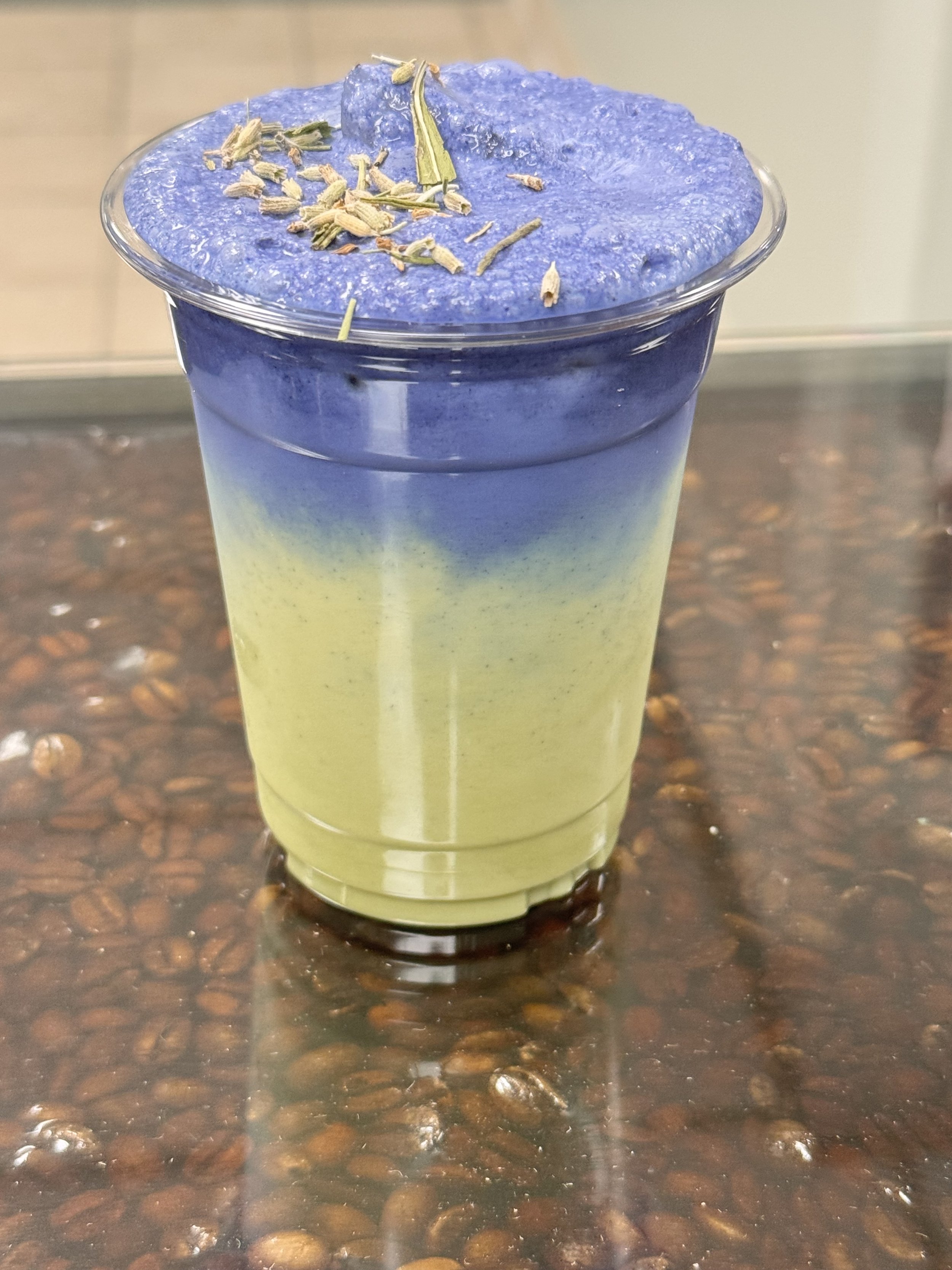 A glass of layered matcha green tea and butterfly pea flower tea topped with dried lavender and sage herbs, on a wooden table with coffee beans.