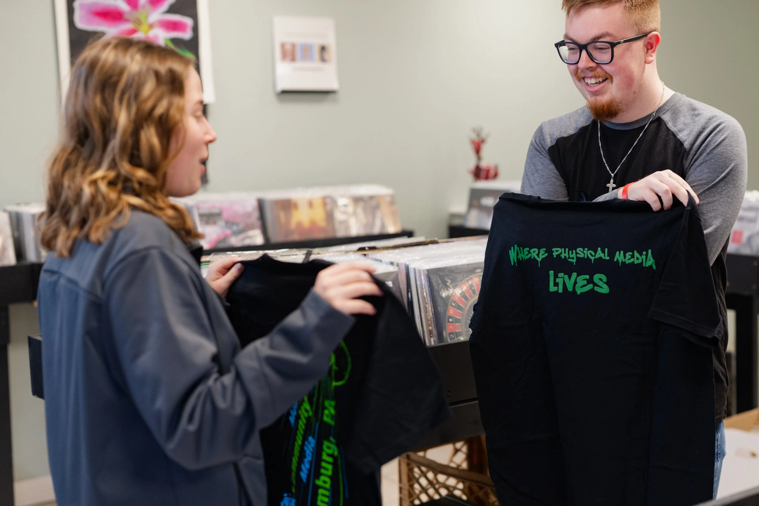 Two people browsing vinyl records in a music store, one is a woman with reddish-brown hair and a gray jacket, the other is a man with glasses, a beard, and a necklace, holding a black bag with green writing. The background shows posters and a display