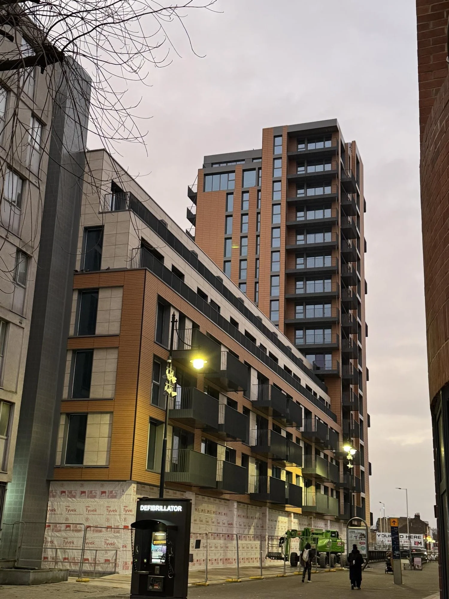 A tall modern apartment building with terrace balconies on each floor. There are streetlights, a defibrillator station, and people walking on the sidewalk, with a green construction vehicle nearby and cloudy sky.