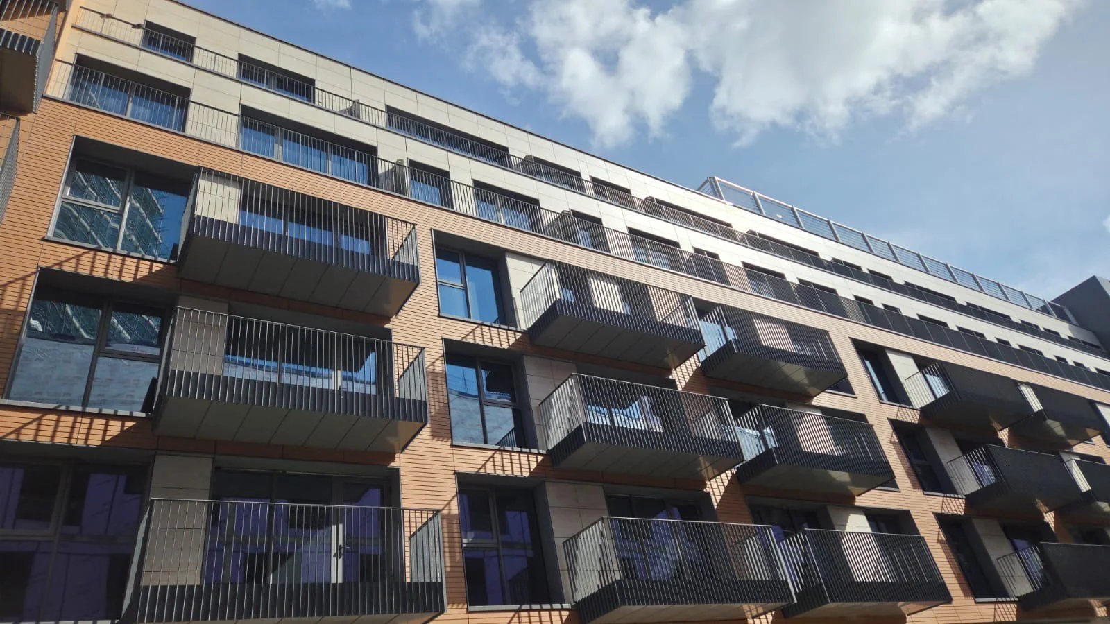 View of a modern multi-story apartment building with balconies and glass windows against a blue sky with scattered clouds.