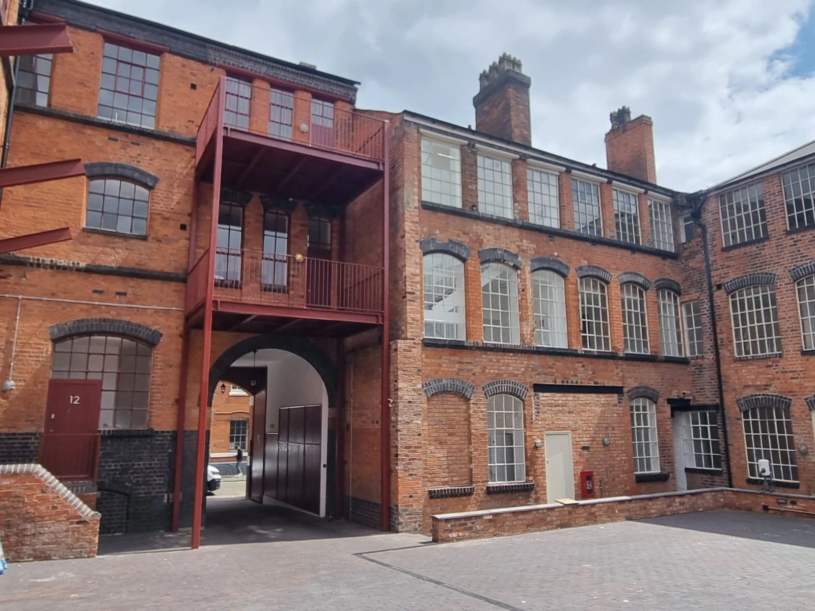 A brick apartment building with multiple floors, large windows, and fire escapes, with an archway entrance leading to a courtyard or alley, under a cloudy sky.