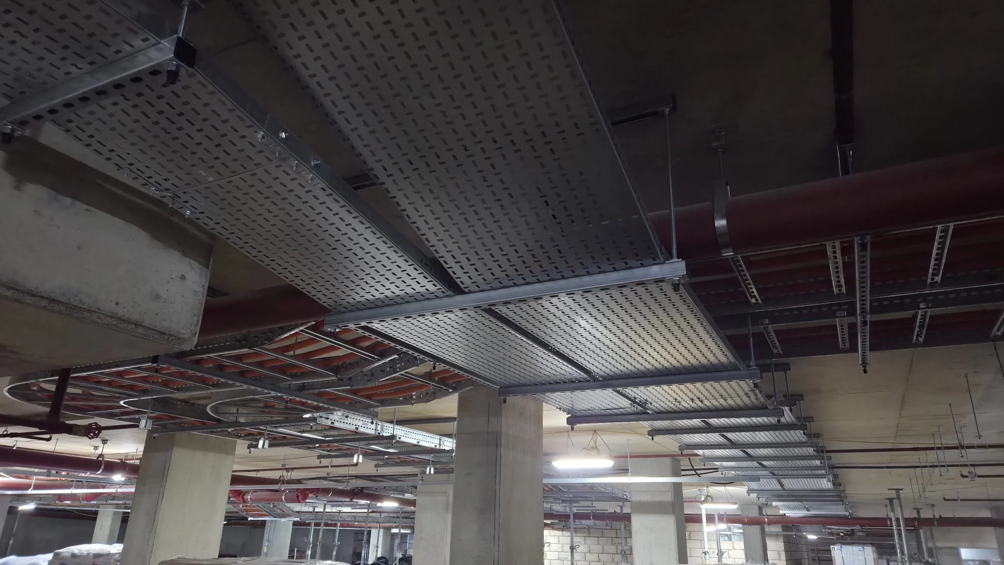 Ceiling of a building with visible metal cable trays, electrical conduits, and pipes under construction.