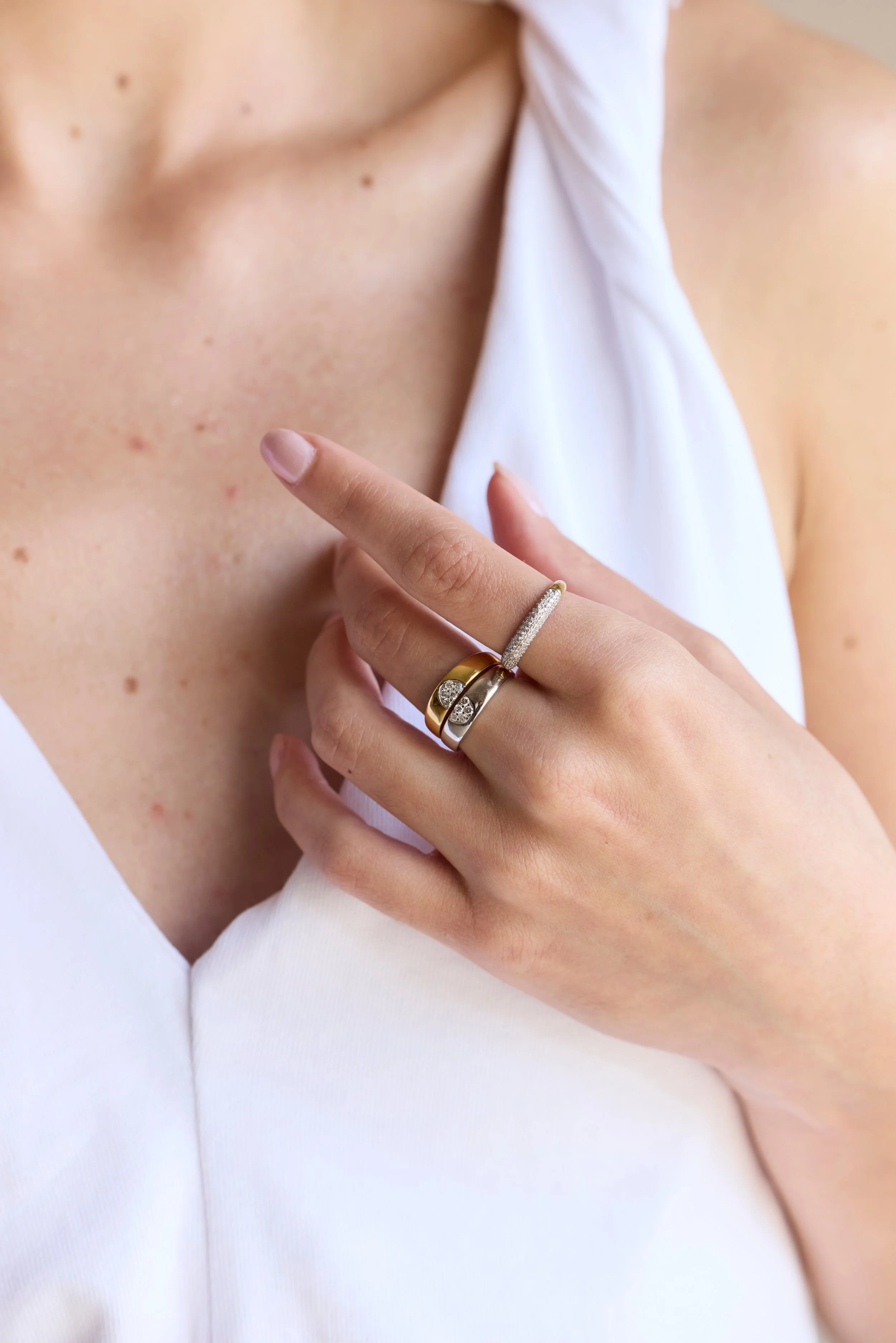 Close-up of a woman's hand adorned with rings, resting on her chest, with visible skin and a portion of a white top.