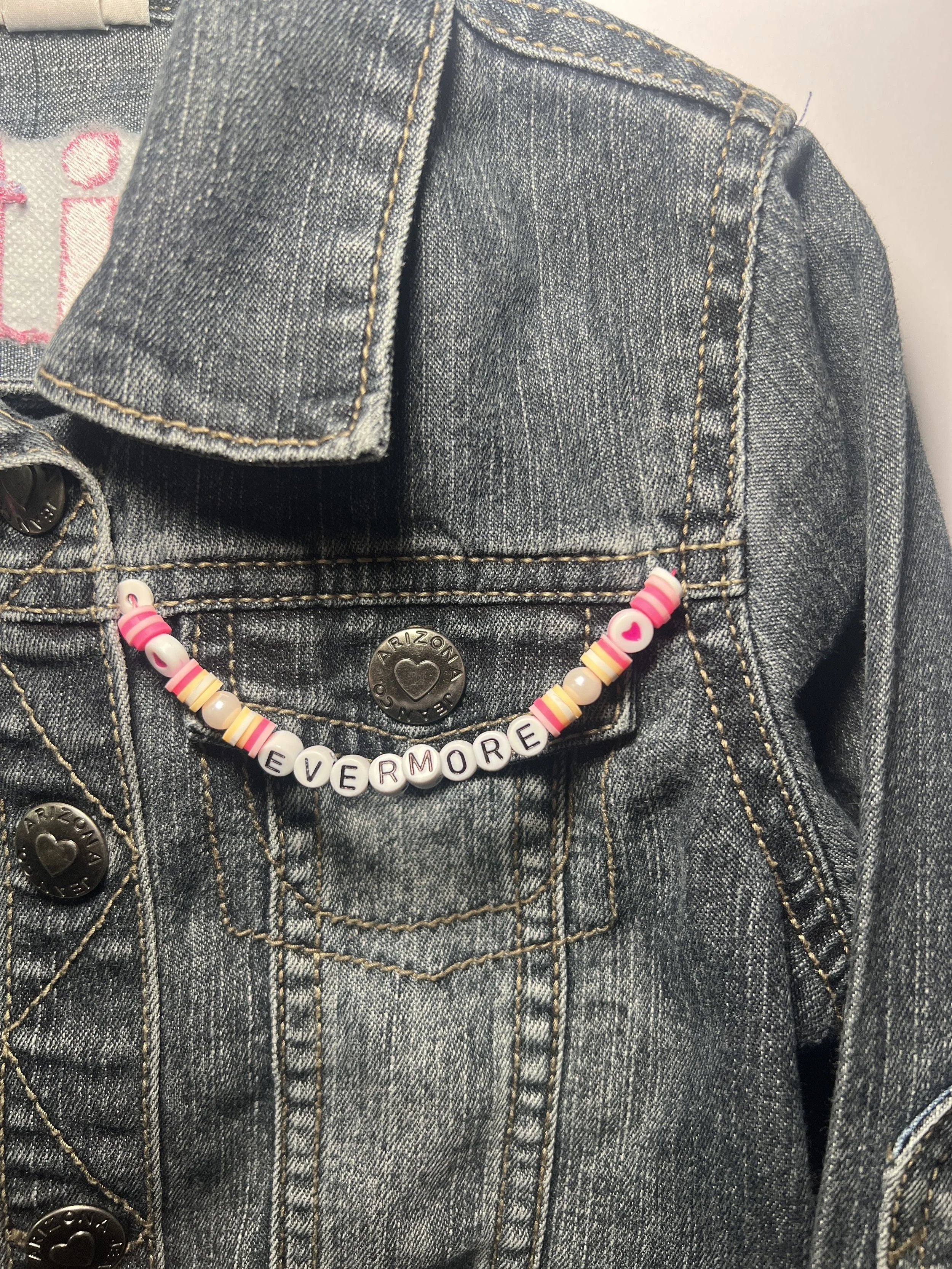 Denim jacket with beaded bracelet spelling 'Evermore' attached to pocket.