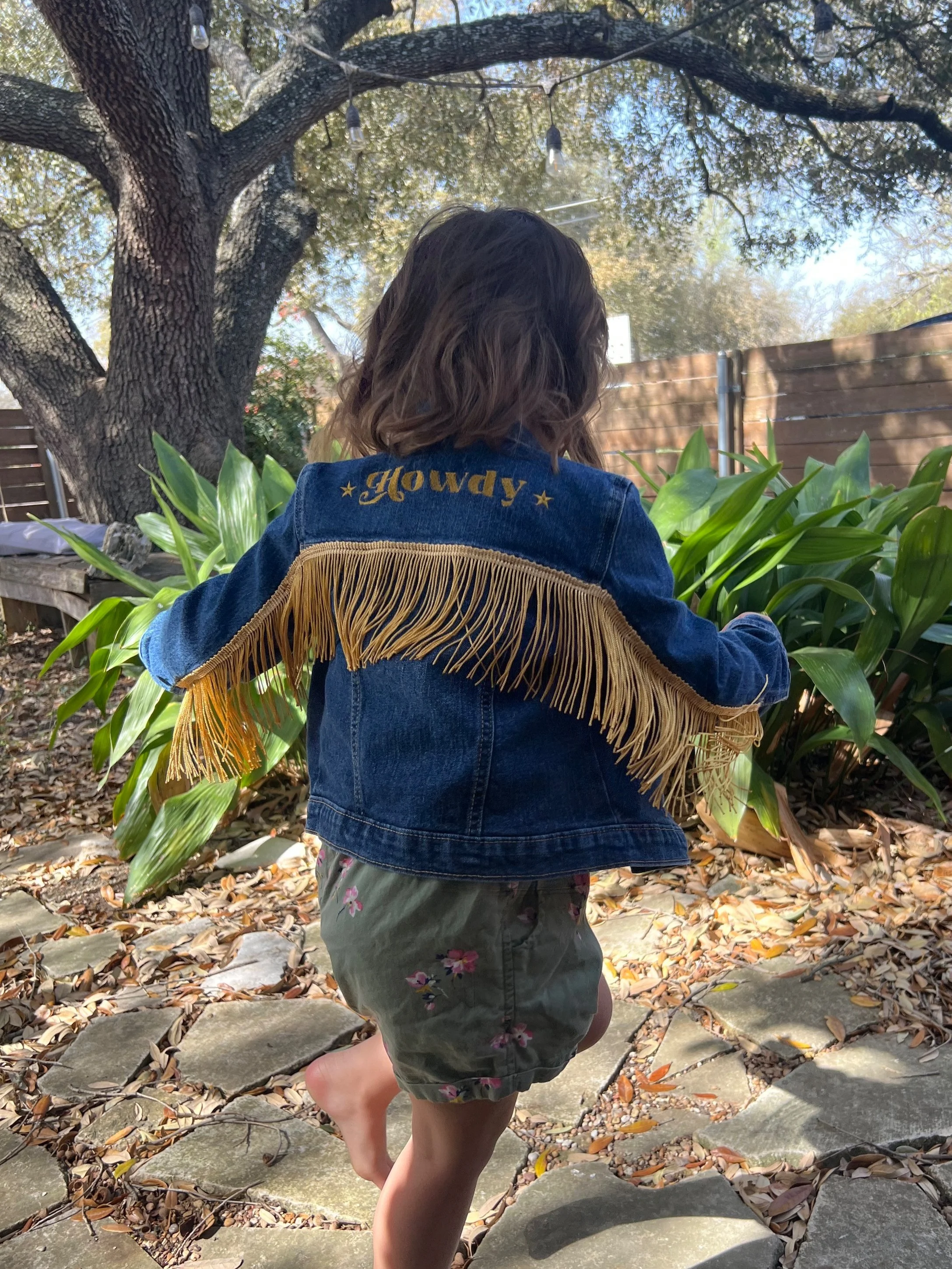 Child wearing denim jacket with "Howdy" and fringe, walking on stone path in garden