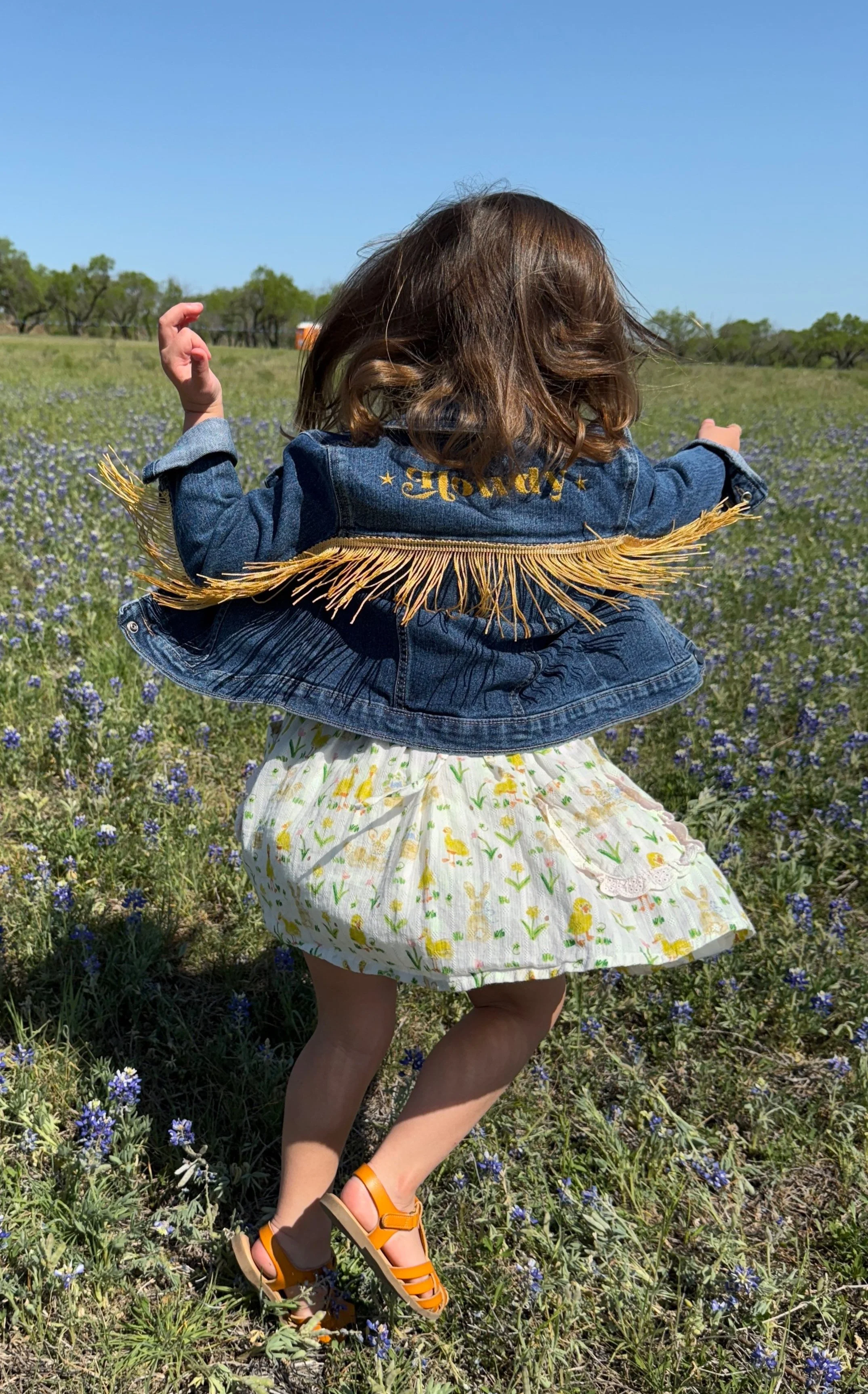 Child in denim jacket with fringe and floral dress dancing in a field of bluebonnets.