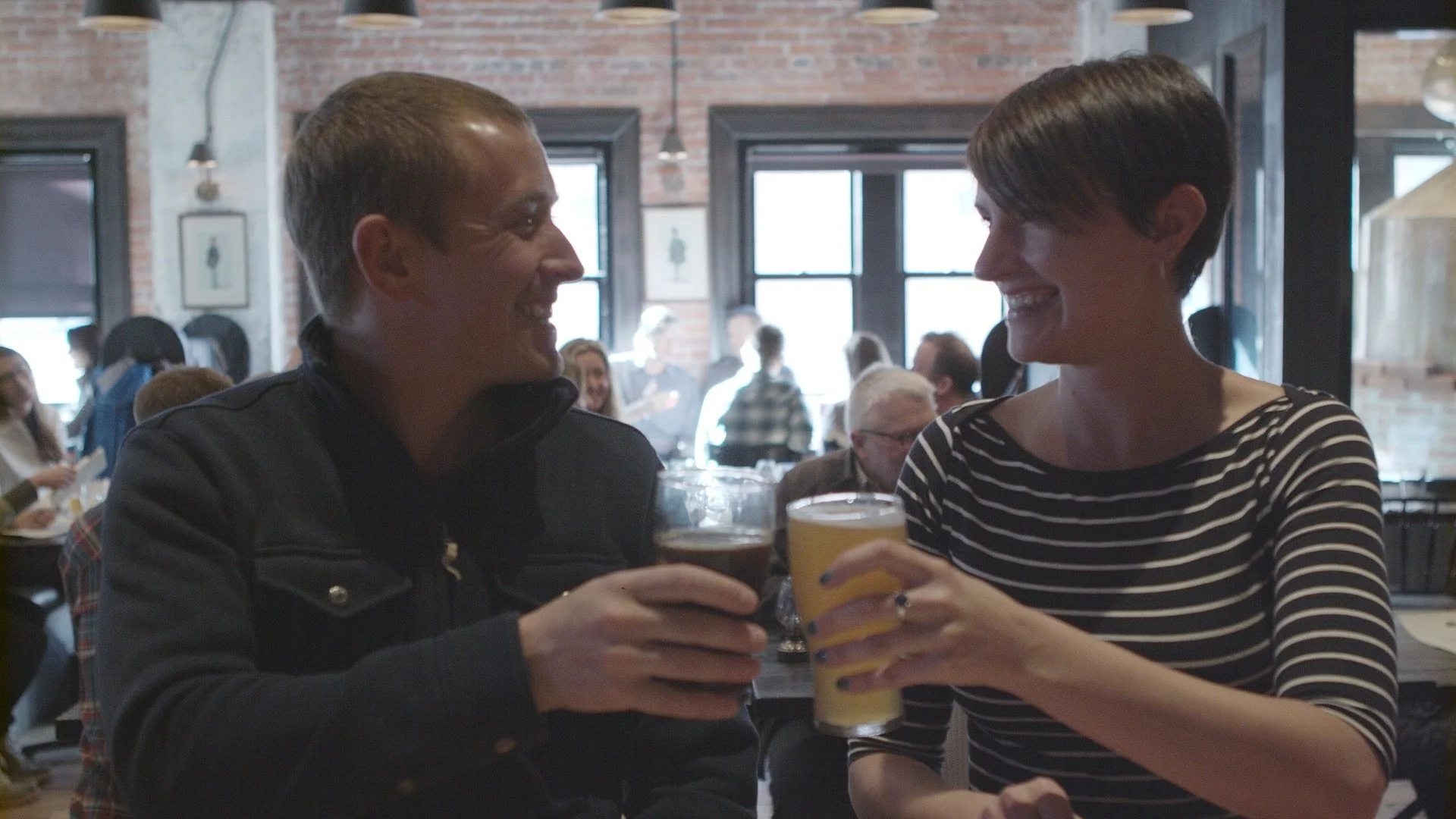 Two people, a man and a woman, smiling and holding drinks, sharing a moment at a lively bar or restaurant.