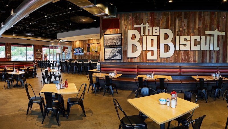 Interior dining room of The Big Biscuit restaurant featuring rustic wood wall branding, booth seating, industrial lighting, and casual breakfast dining tables.