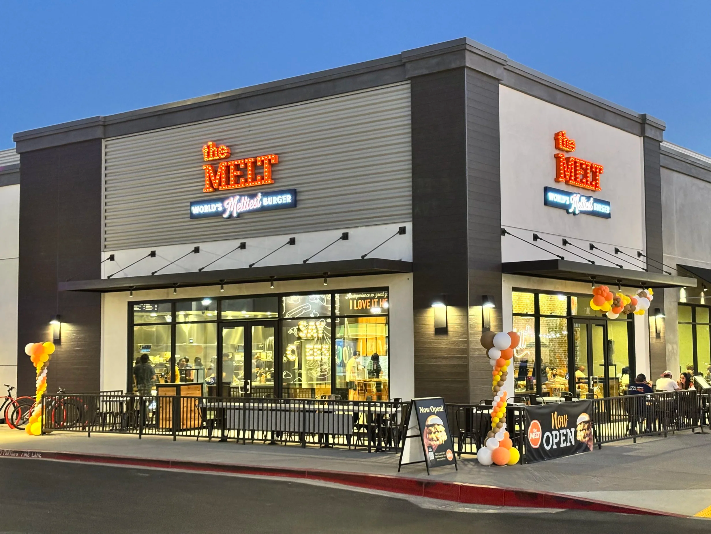 Exterior of The Melt restaurant with illuminated signage, outdoor patio seating and customers dining inside.