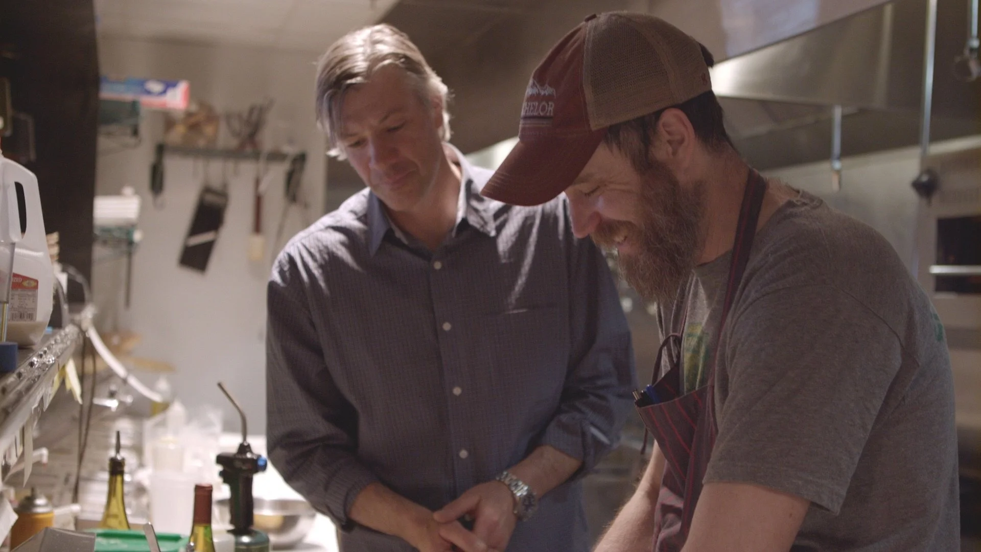 Two men in a kitchen, one with gray hair and the other with a beard and cap, appear to be preparing or working on something together, smiling and focused.