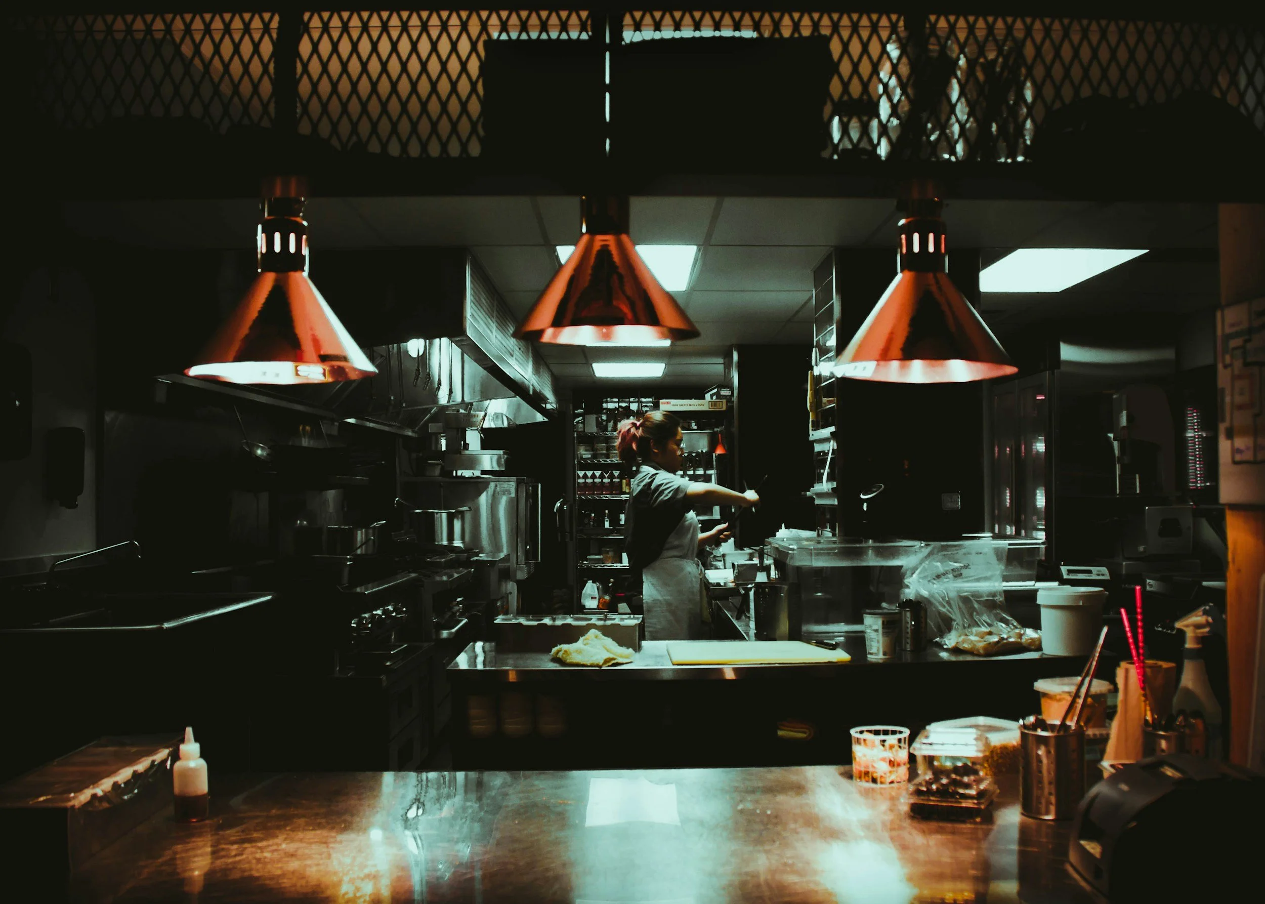 A restaurant kitchen with a worker preparing food, illuminated by red pendant lights, with various cooking utensils and ingredients on the countertop.