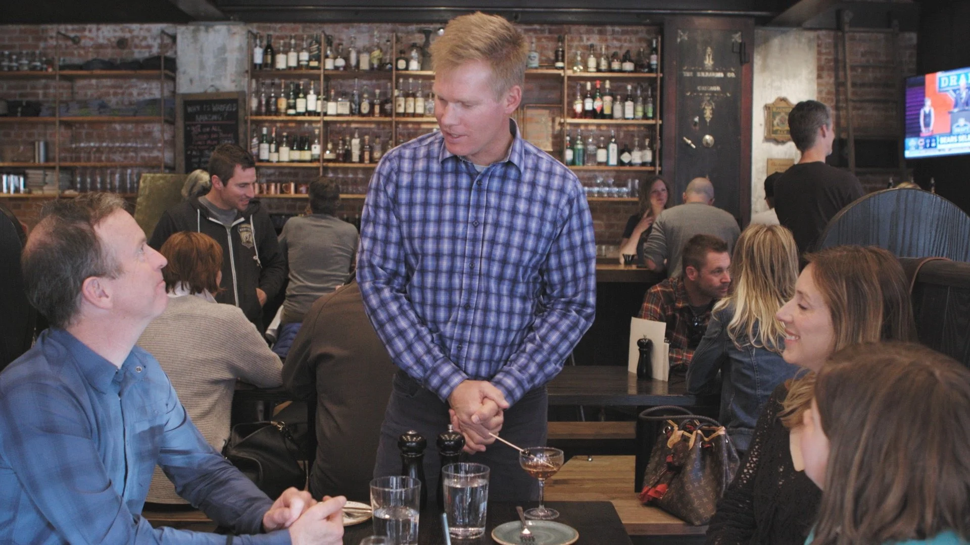 A man in a blue checkered shirt is standing and smiling at a woman seated at a table in a busy restaurant or bar, with several other patrons sitting and talking at their tables in the background. The setting has a brick wall with shelves of bottles behind the bar.