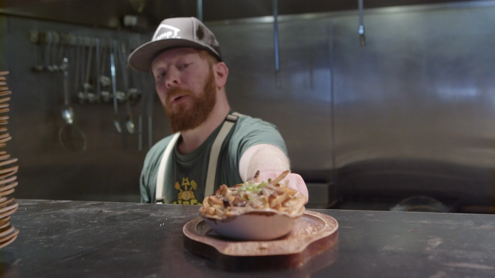 A man with a beard and wearing a cap and apron offers a slice of pizza topped with chicken and vegetables in a kitchen setting.