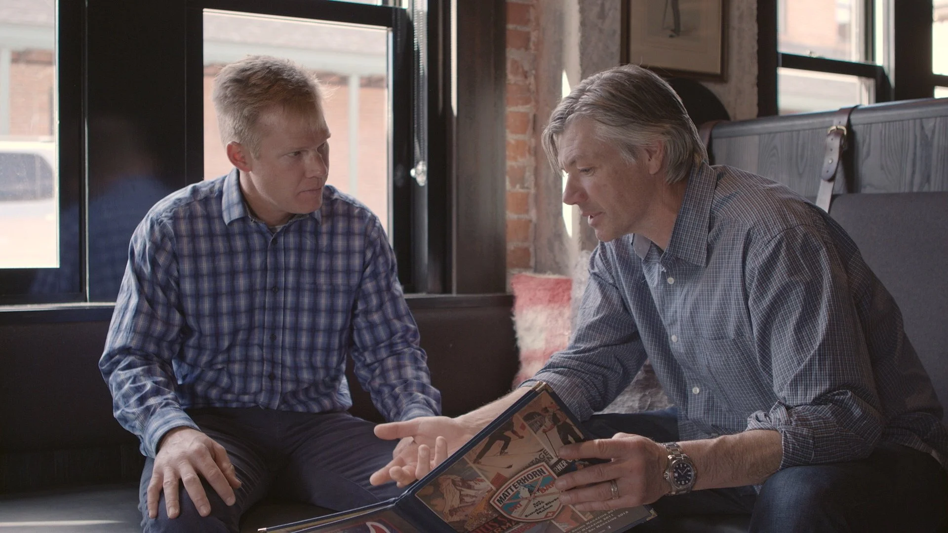 Two men sitting indoors with exposed brick walls and large windows, looking at a comic book.