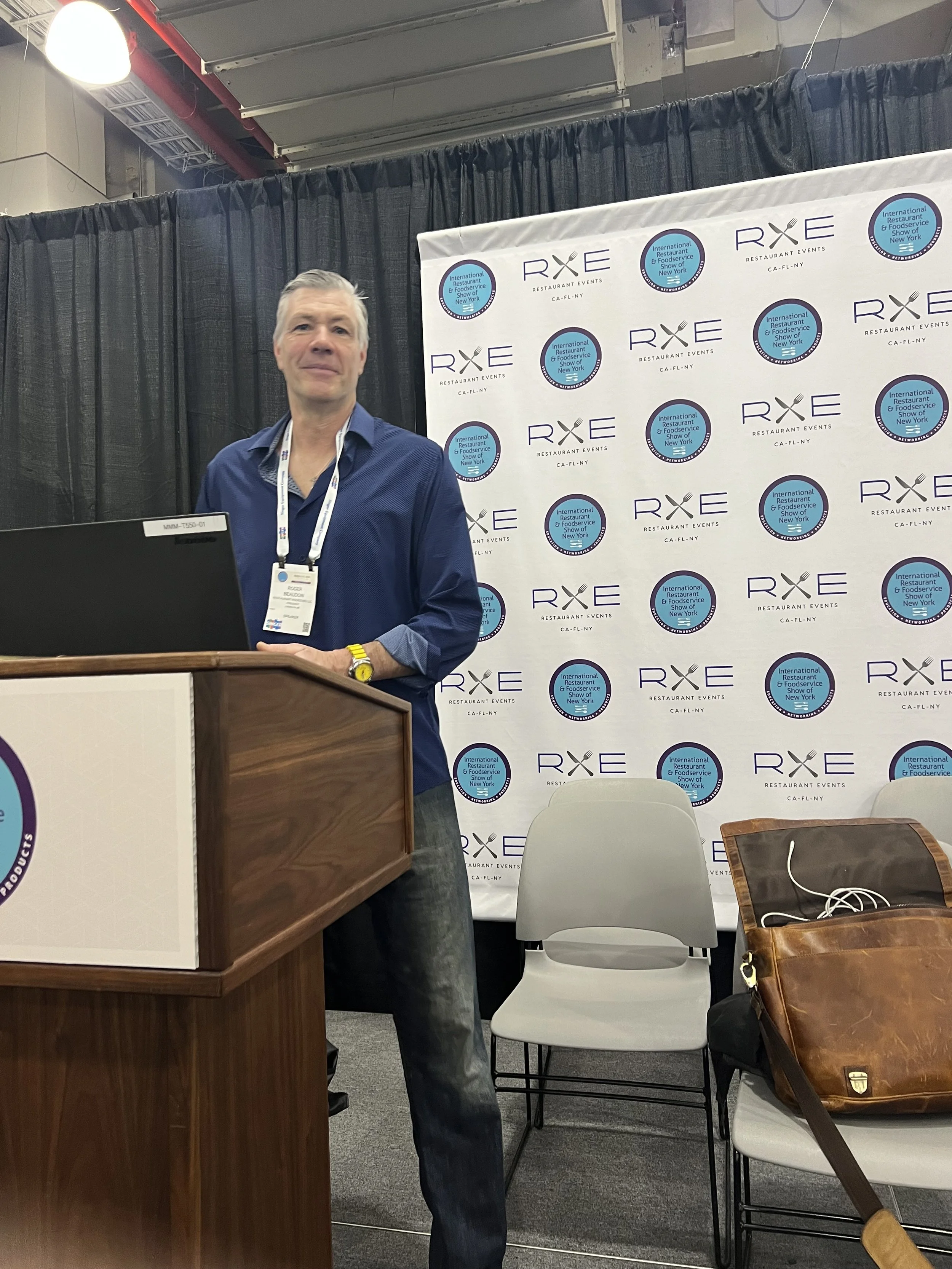 A man with gray hair, wearing a blue shirt and a yellow watch, stands at a wooden podium in front of a backdrop with logos that read ROI Restaurant Events and International Restaurant & Foodservice Show of New York. There are black curtains behind the backdrop, and a small table with a brown leather bag and some earphones is in the foreground.