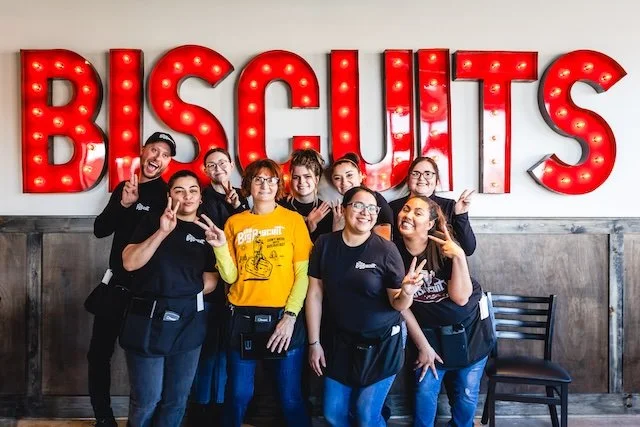 Big Biscuit restaurant team members posing together in front of a large illuminated “Biscuits” wall sign inside the restaurant.