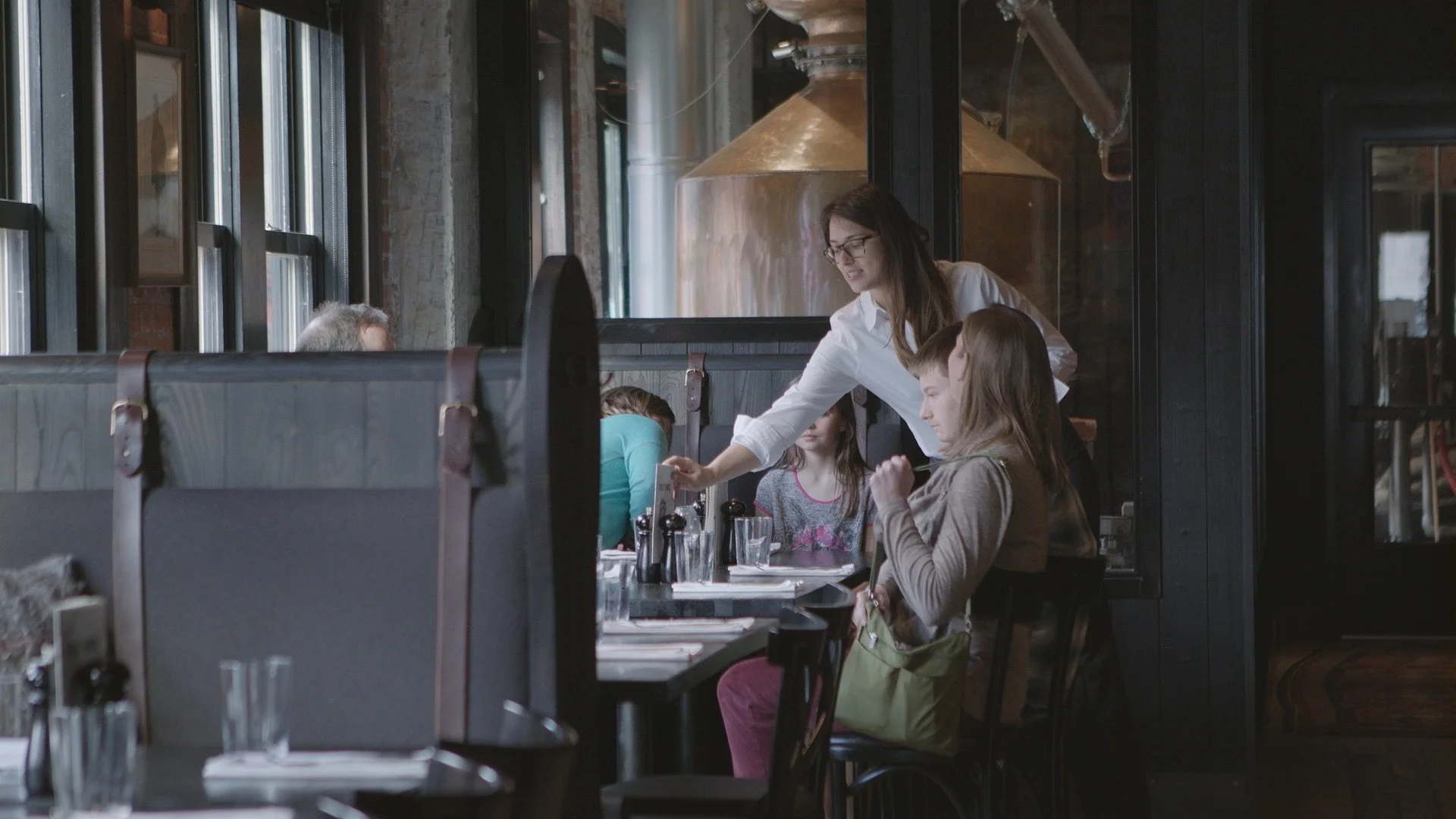 A woman waitress serving drinks to children seated at a table in a modern restaurant with industrial decor, large windows, and exposed brick walls.