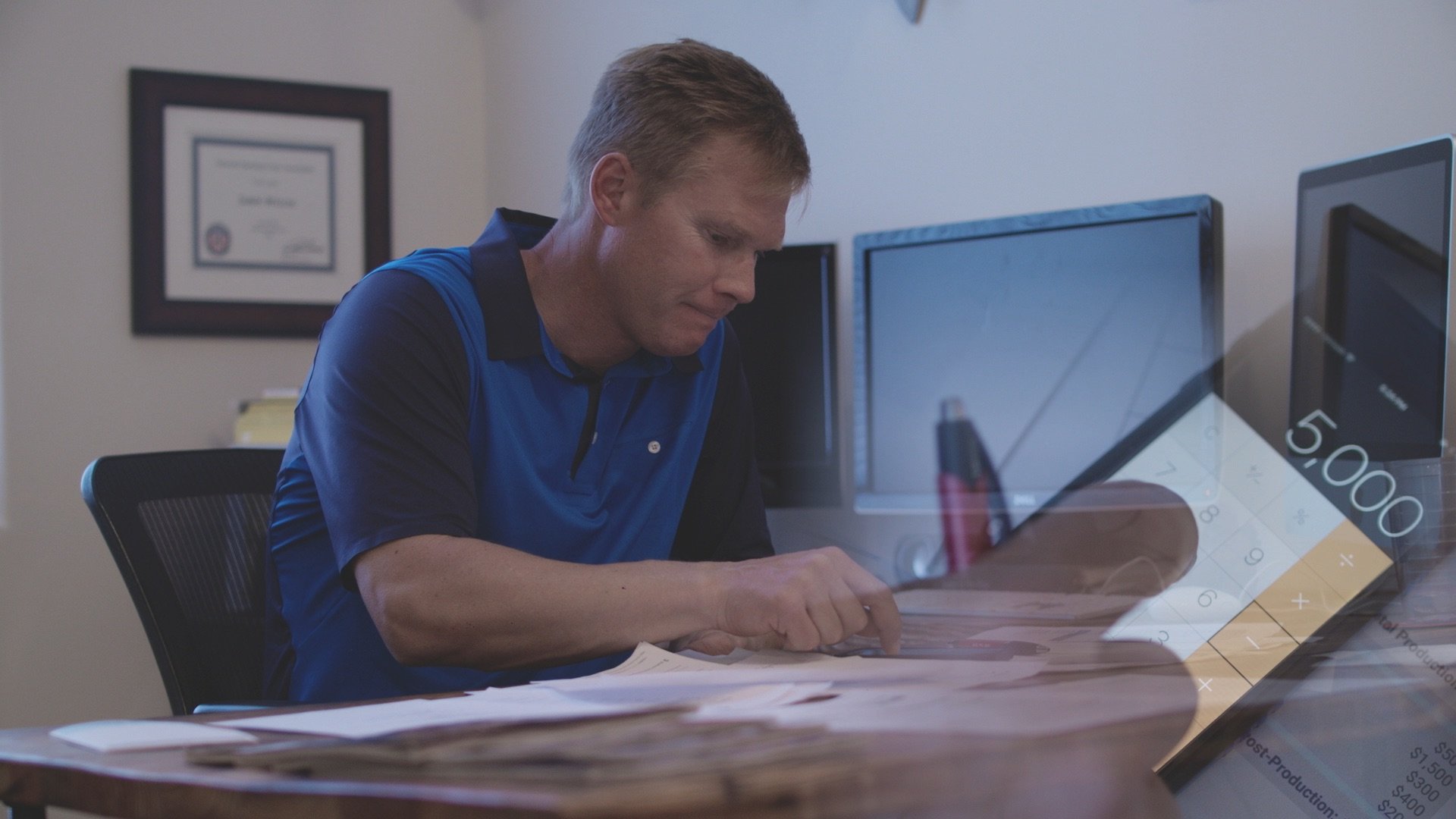 A man in a blue and black polo shirt sitting at a desk, looking down at papers with a focused expression. Multiple computer monitors and a framed certificate are on the wall behind him, with a digital clock showing 5:00 on a transparent overlay.