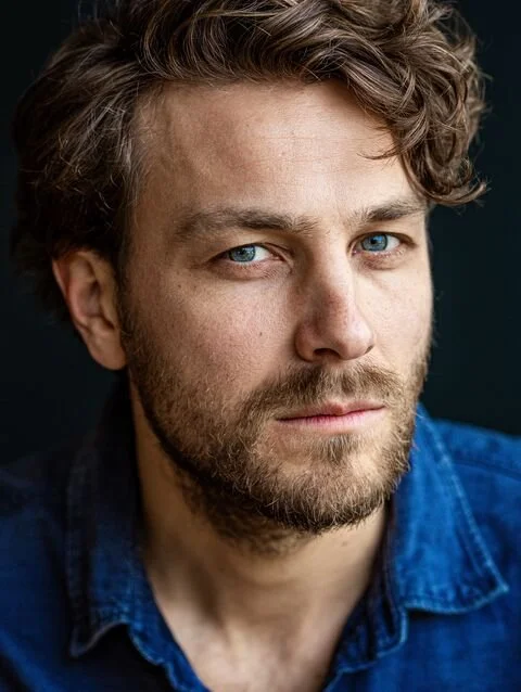 Ein Mann mit braunen Haaren und Bart, in einer schwarzen Lederjacke, schaut in die Kamera. Der Hintergrund ist dunkel