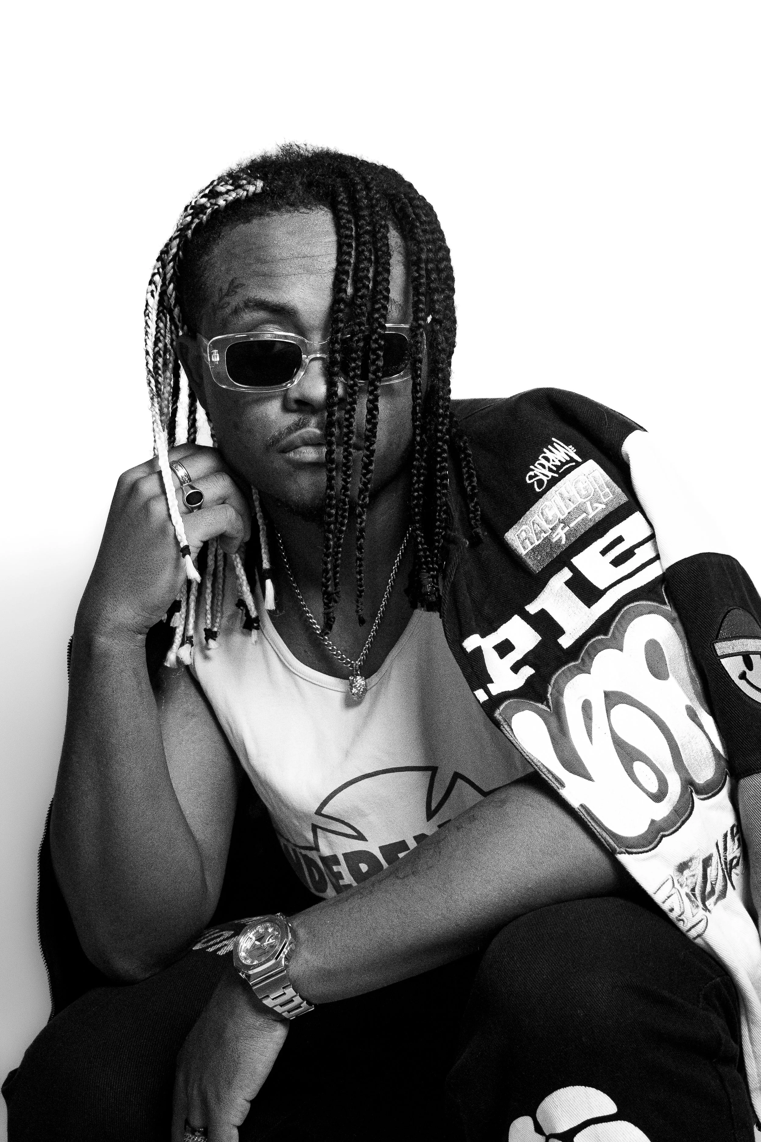 Un homme portant des lunettes de soleil, des chaînes et un gilet sans manches avec des logos, pose de manière décontractée contre un mur blanc.