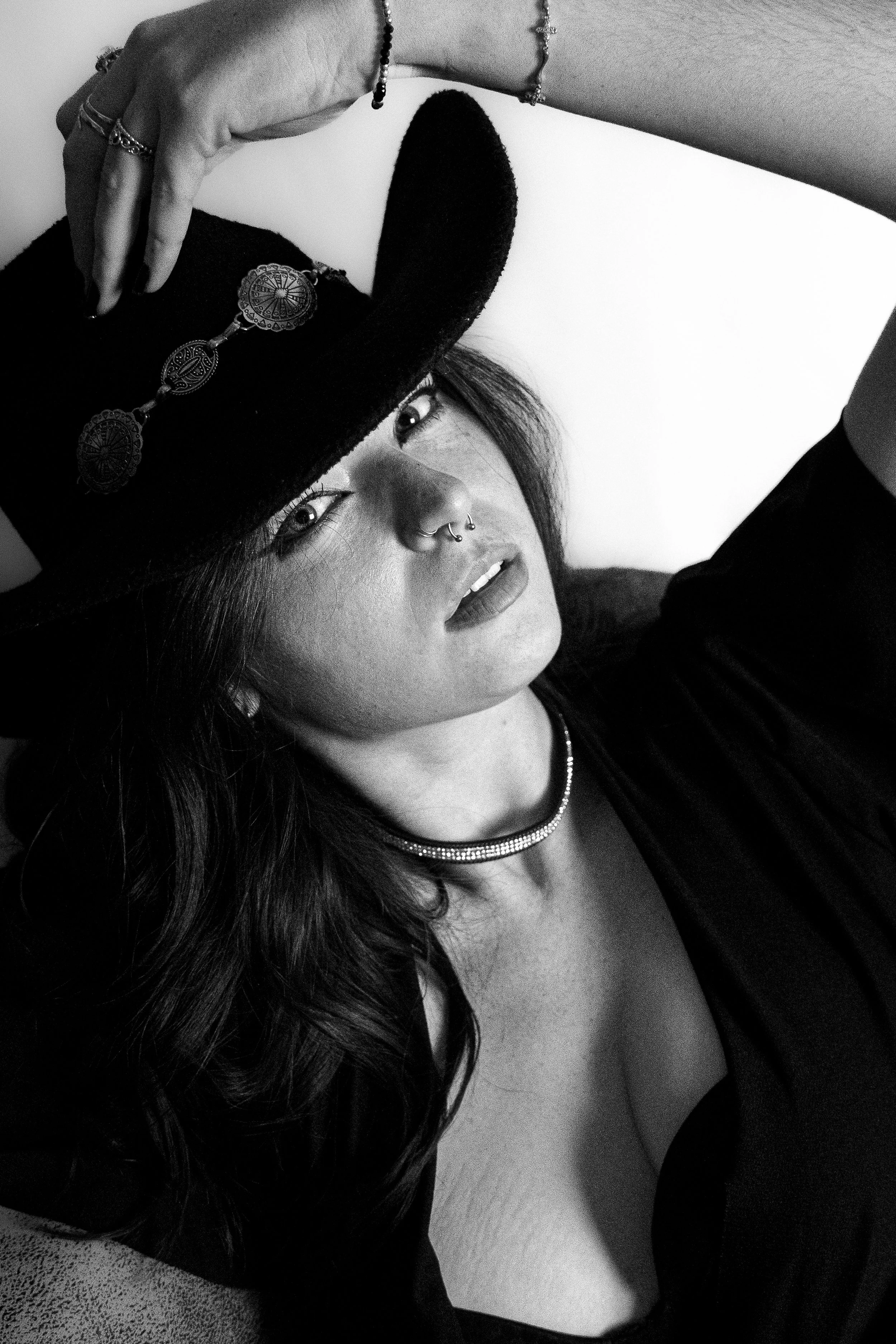 Une femme porte un chapeau noir avec des décorations, un collier et un piercing au nez, elle regarde l'objectif en posant.